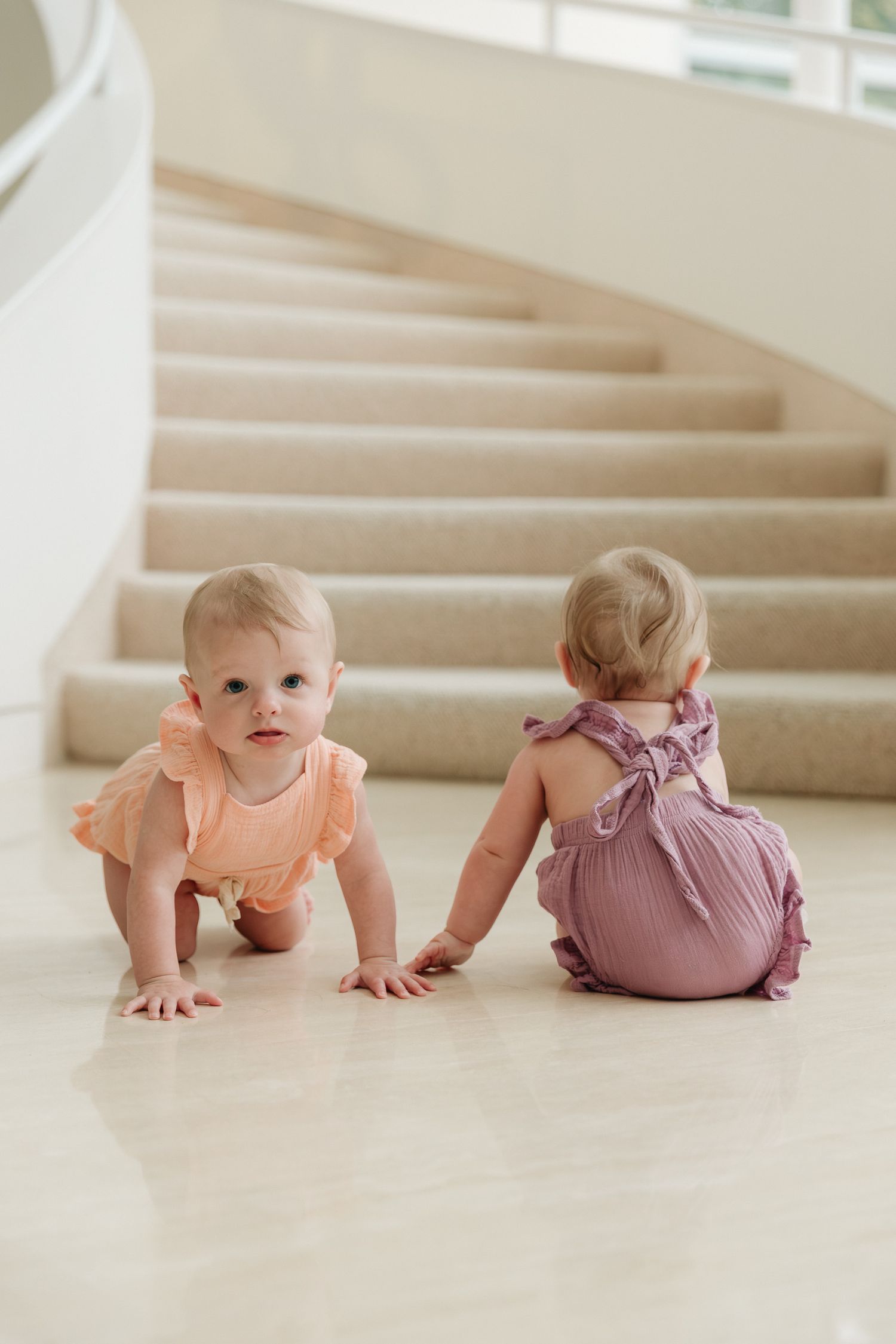Two babies in peach and mauve rompers crawl on a polished marble floor in front of a curved, carpeted staircase.