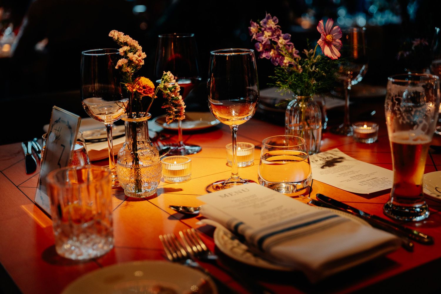 Candlelit restaurant table set with wine glasses, folded napkins, silverware and small flower vases for an intimate dinner.