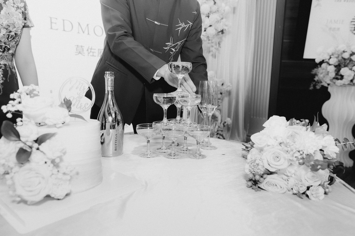 Black and white photo of a person pouring champagne into a stacked glass tower beside a tiered wedding cake adorned with white roses.