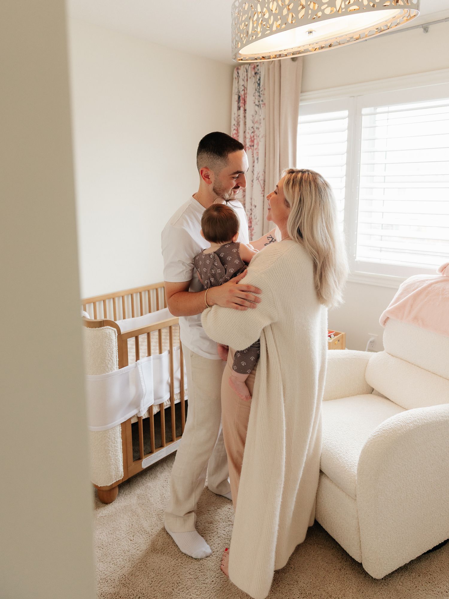 Smiling couple in cream loungewear holding their baby beside a wooden crib in a bright nursery with a cozy white armchair.