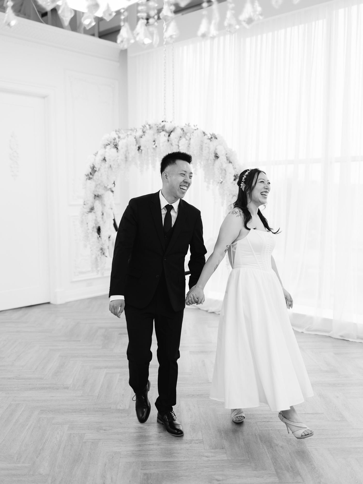Laughing bride in a white gown and groom in a dark suit hold hands while walking past a floral wedding arch in a bright venue.