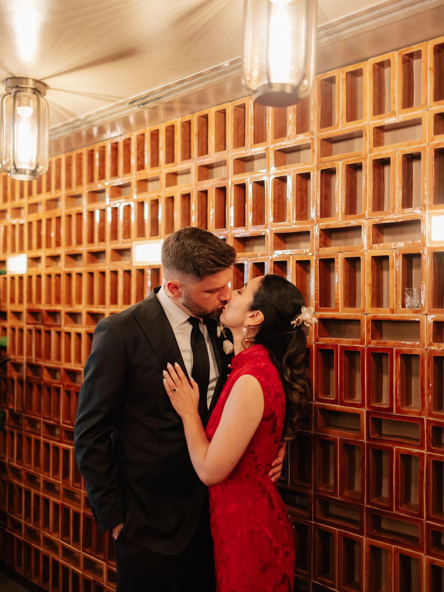A man in a dark suit kisses a woman in a red lace dress in front of a warm terracotta brick lattice wall with hanging lanterns.