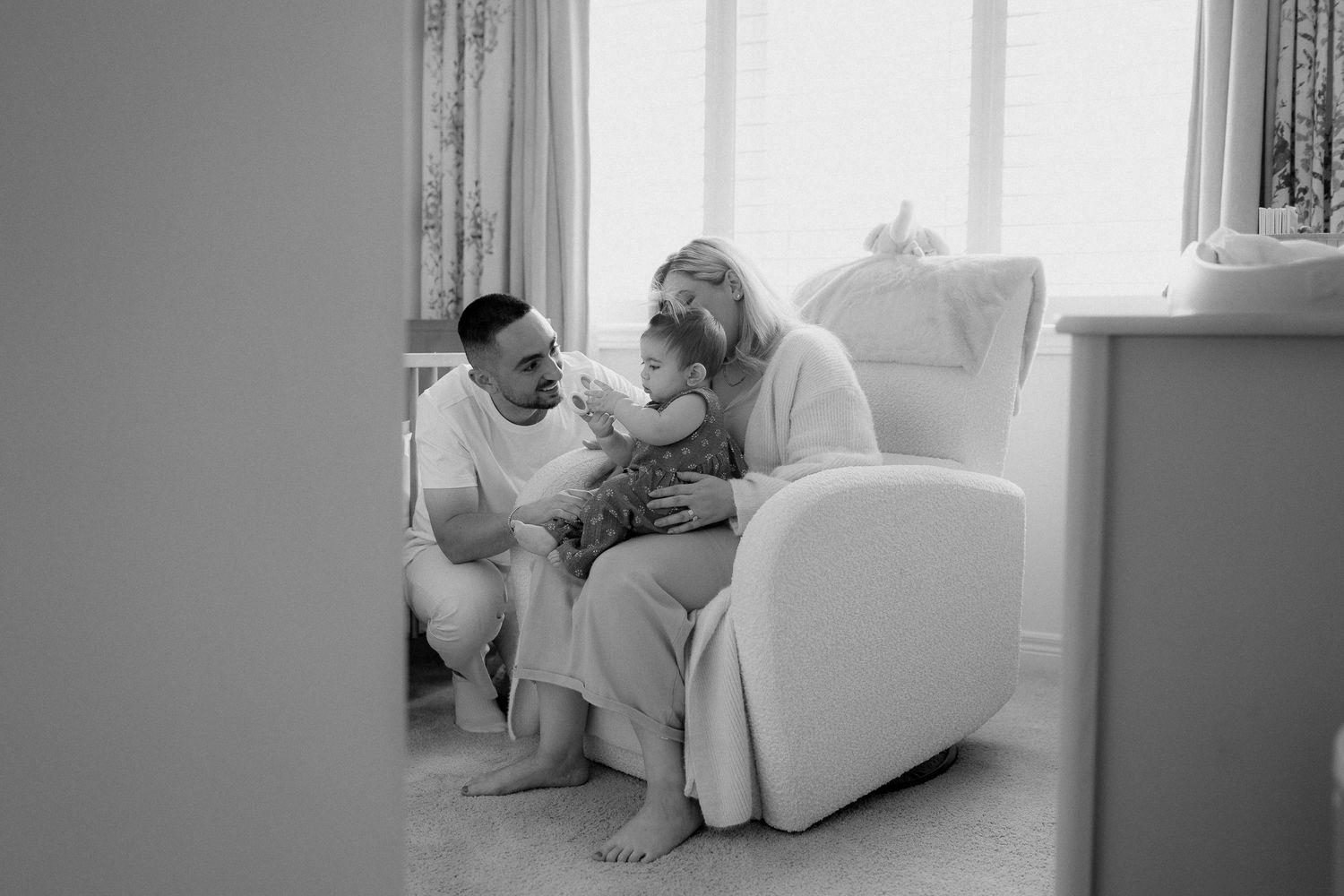 Black and white lifestyle photo of parents sitting with their baby in a cozy nursery beside a white armchair and sunlit window.