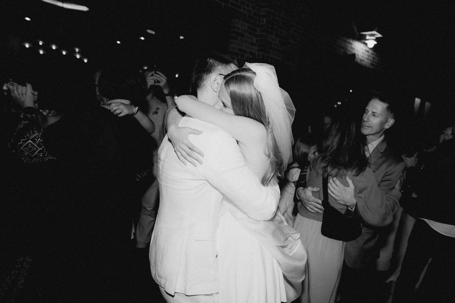 Black and white photo of a bride in a veil and groom in a light suit embracing on a crowded dance floor at their wedding reception.