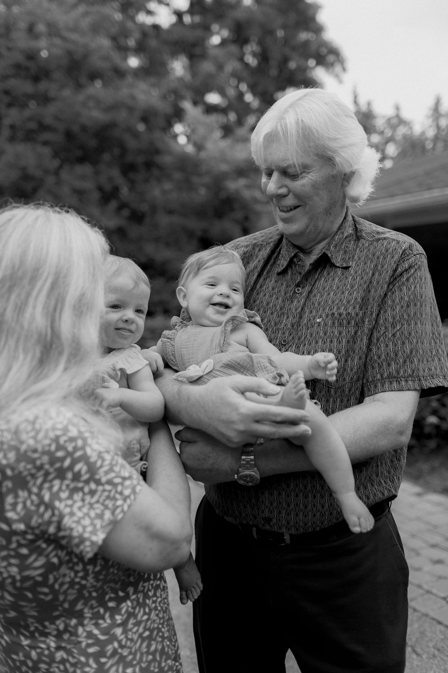 Black and white photo of a smiling grandfather holding a laughing baby while a grandmother holds another toddler outdoors.