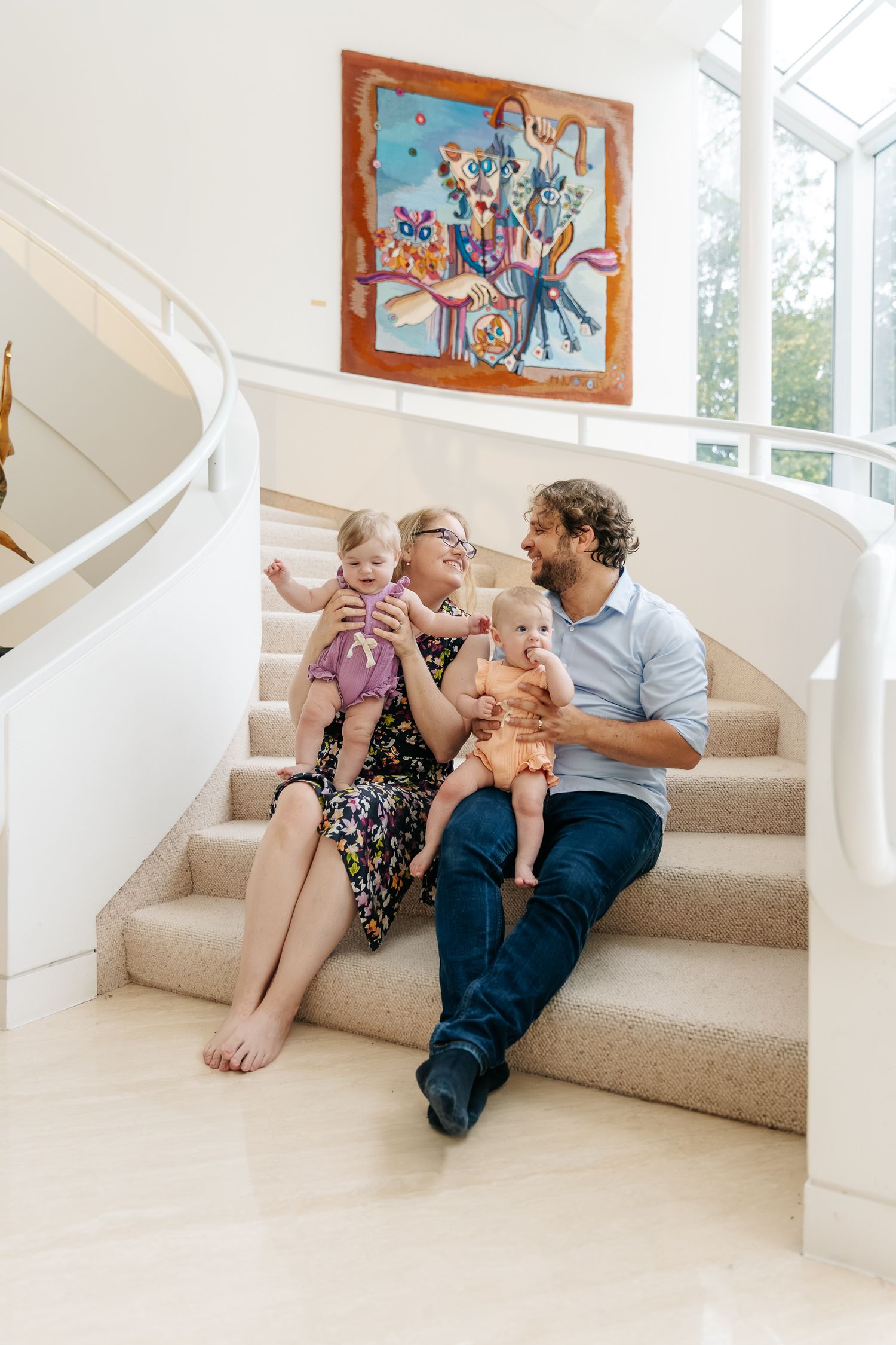 Smiling family of four with two babies sitting on a carpeted curved staircase beneath a colorful abstract painting in a bright modern home.
