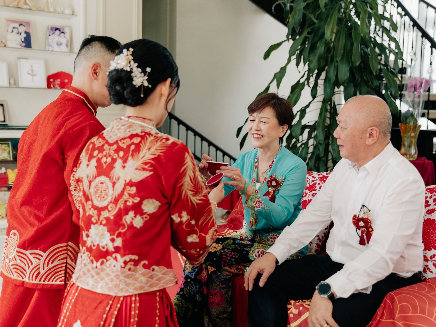 Bride and groom in red traditional Chinese wedding attire share a tea ceremony moment with two smiling parents seated in a bright room.