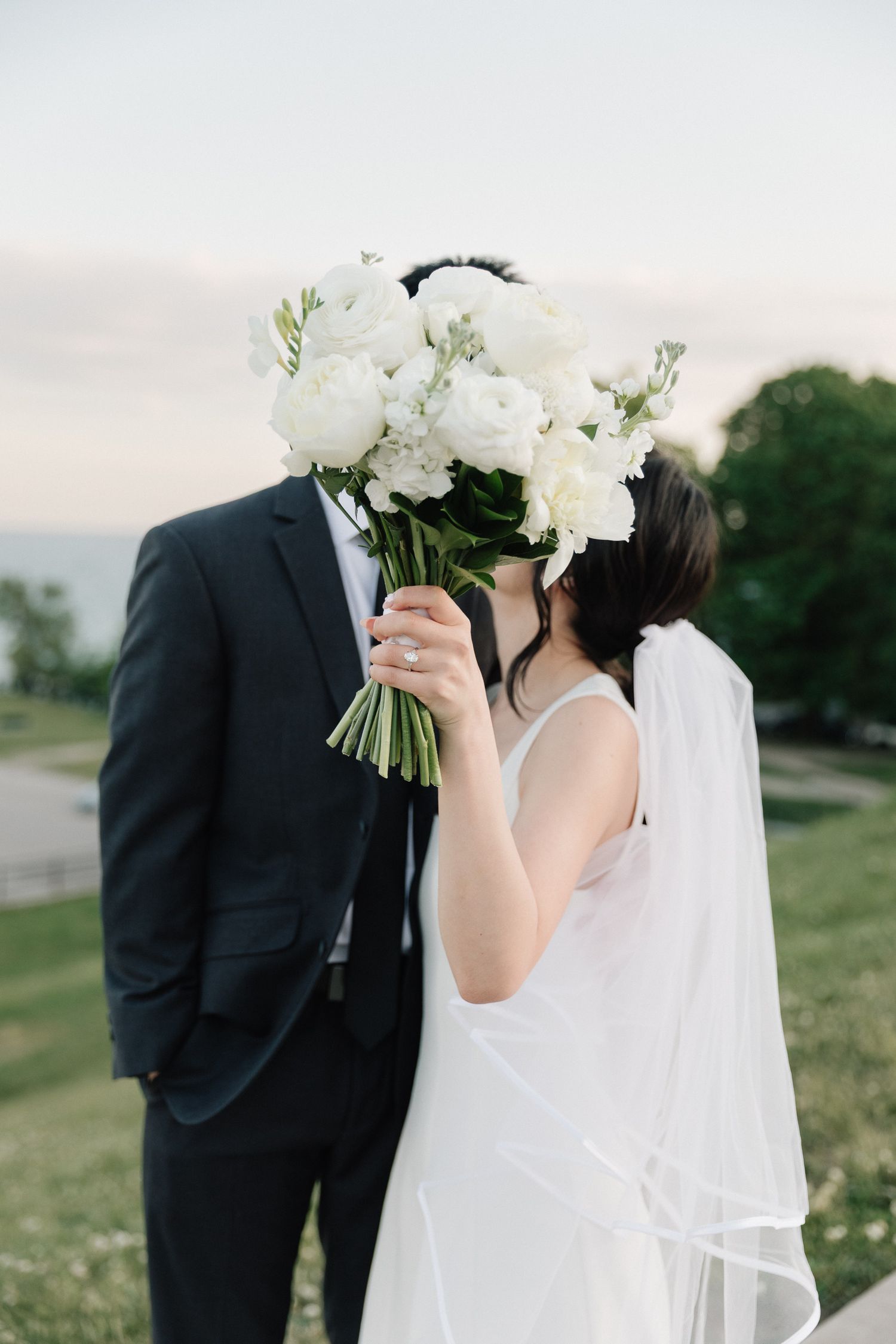 Bride and groom kissing behind a white floral bouquet outdoors, bride wearing a white gown and veil.