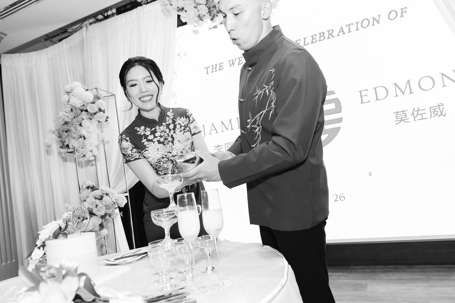 A smiling bride in a floral cheongsam and a groom in a dark shirt pour champagne into glasses at a wedding celebration table.