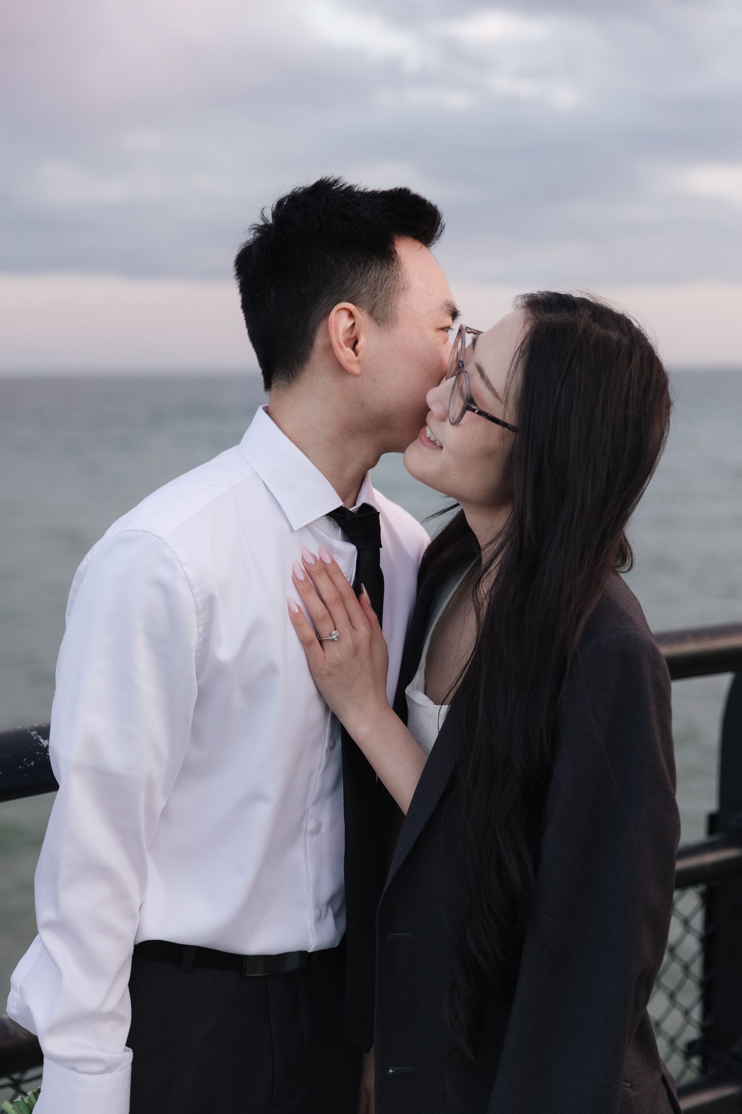 Man in white shirt and tie kisses the cheek of a smiling woman wearing glasses and a blazer, her hand on his chest, by the ocean at dusk.