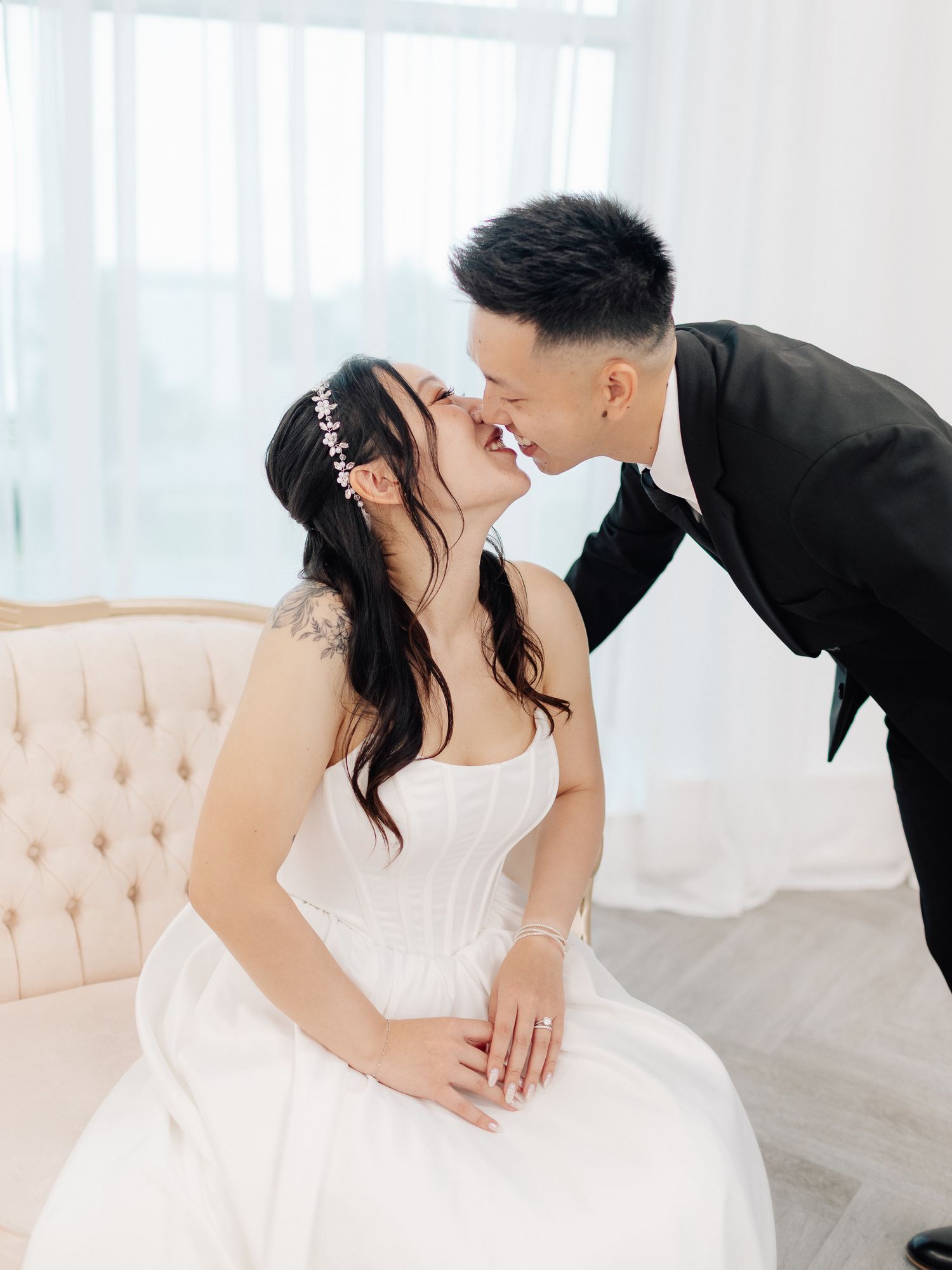 Bride in a white gown with a floral hair accessory shares a kiss with her groom in a black suit on a tufted cream sofa.