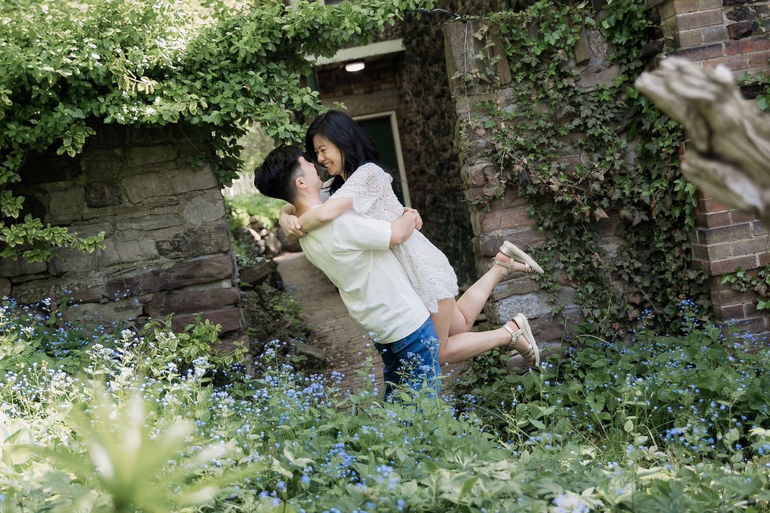 Couple embracing and lifting one another in a lush garden with wildflowers, ivy-covered stone walls, and brick in the background.