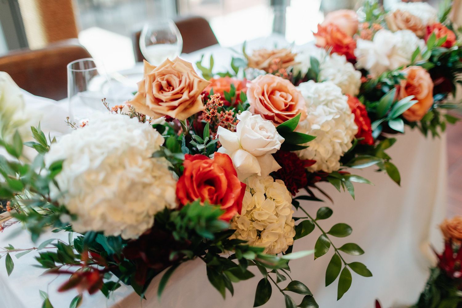 Wedding table centerpiece of cream, coral and orange roses with white hydrangeas and green foliage on a white-linen table with wine glasses.