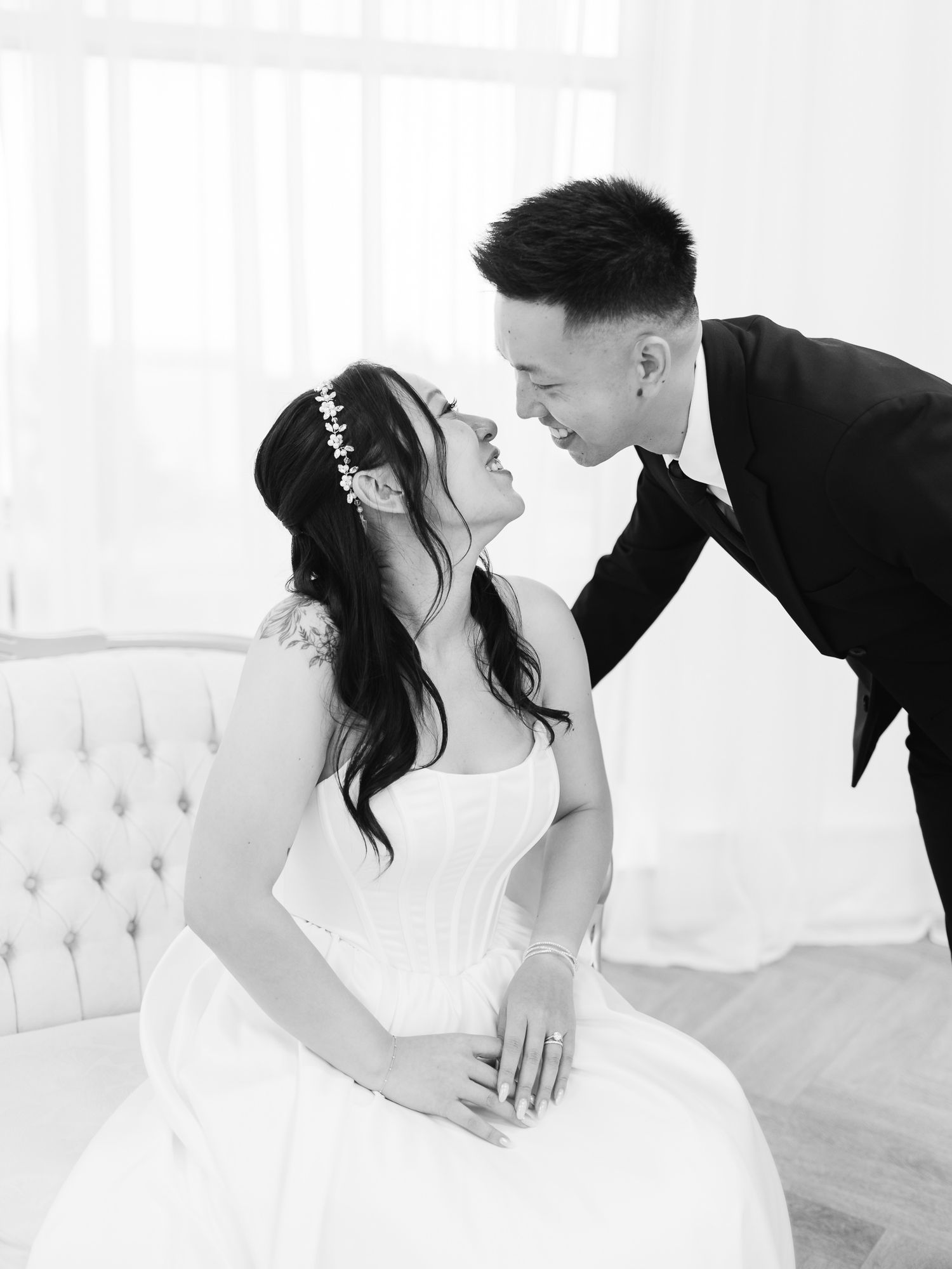 Black and white photo of a bride in a strapless gown and floral headpiece smiling at her groom as he leans in close.