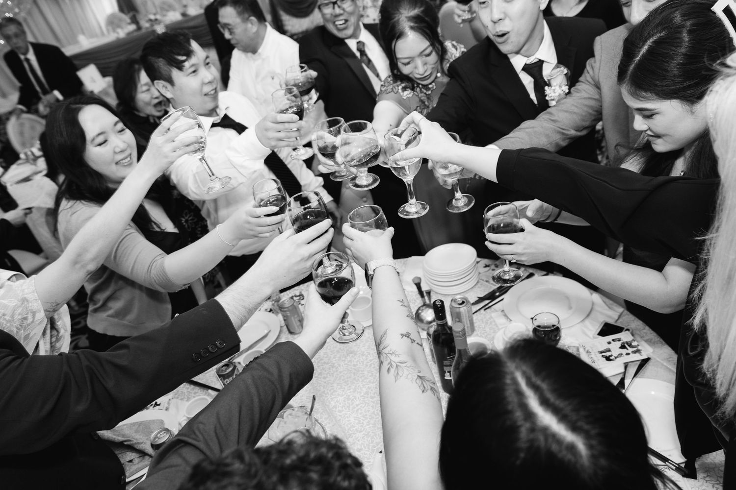 Black and white photo of wedding guests raising glasses in a lively toast around a banquet table, smiling and clinking drinks together.