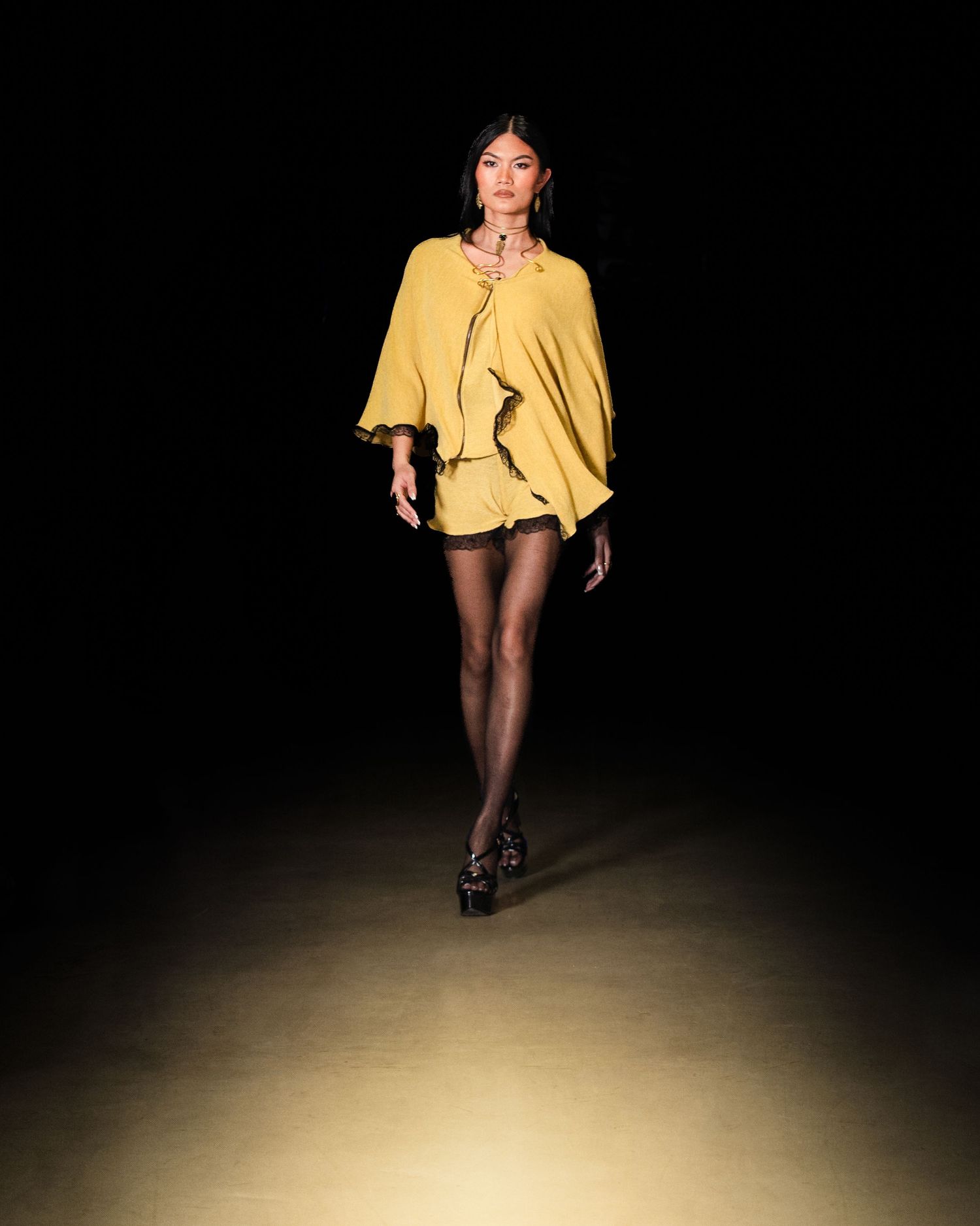 Model walks runway in mustard yellow ruffled cape mini dress with sheer black tights and platform heels.
