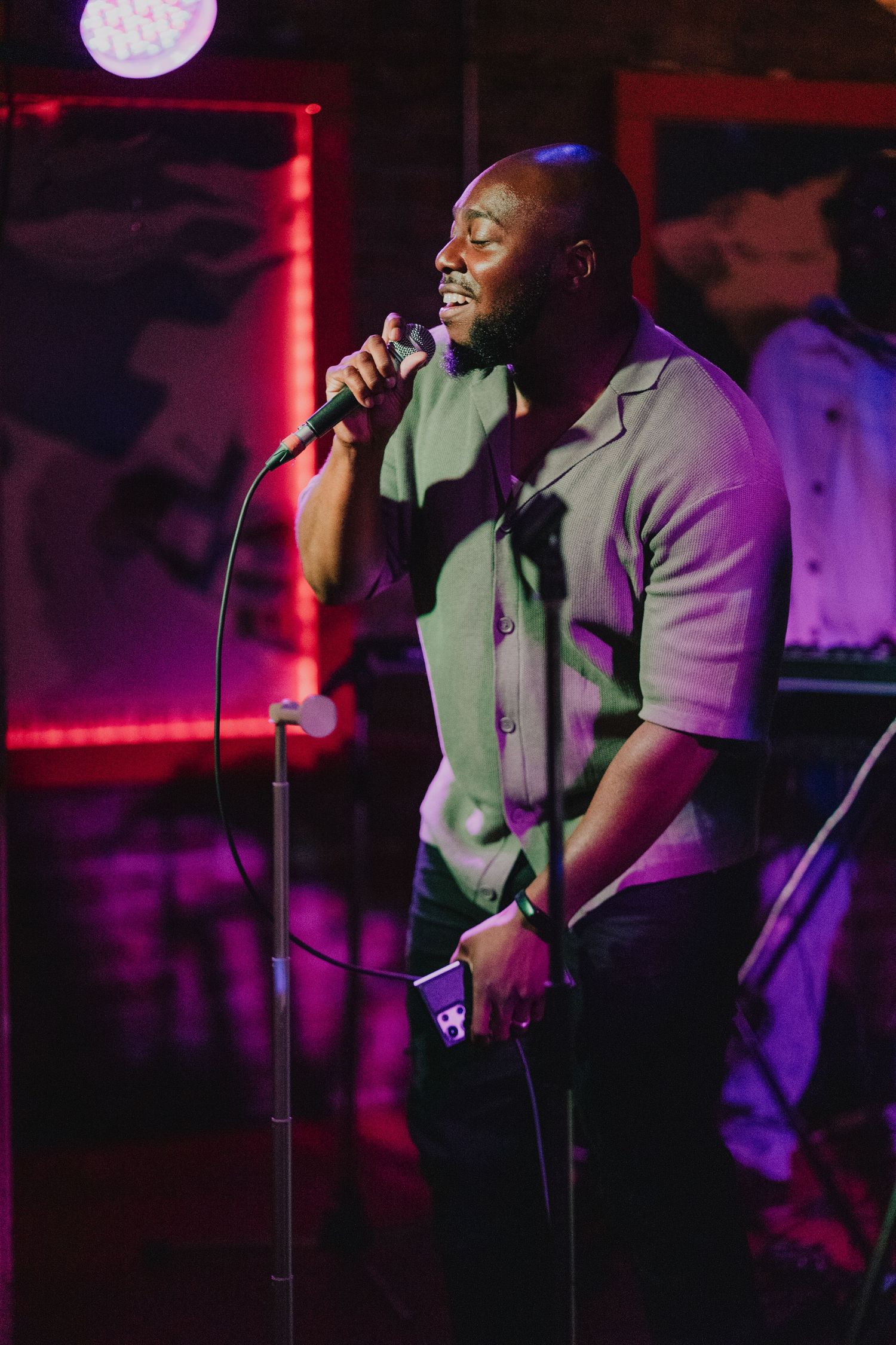 A Black male singer performs passionately on stage with a microphone stand under purple stage lighting.