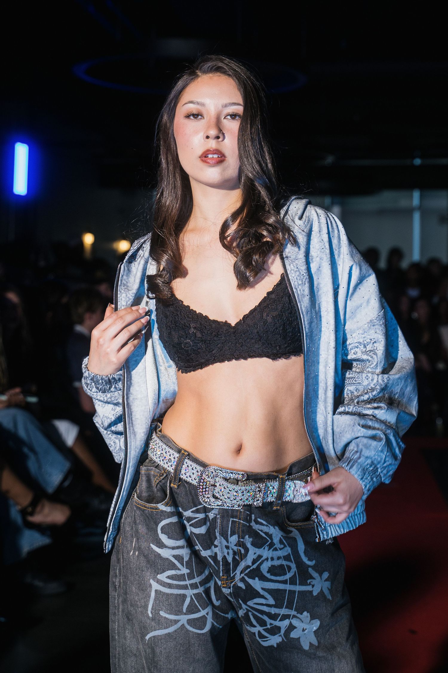 Female model walks runway in silver denim jacket with black lace bralette and graffiti print pants.