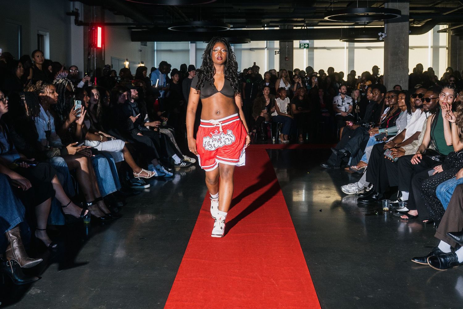 Model walks red carpet in red sports bra and matching shorts at urban fashion show.