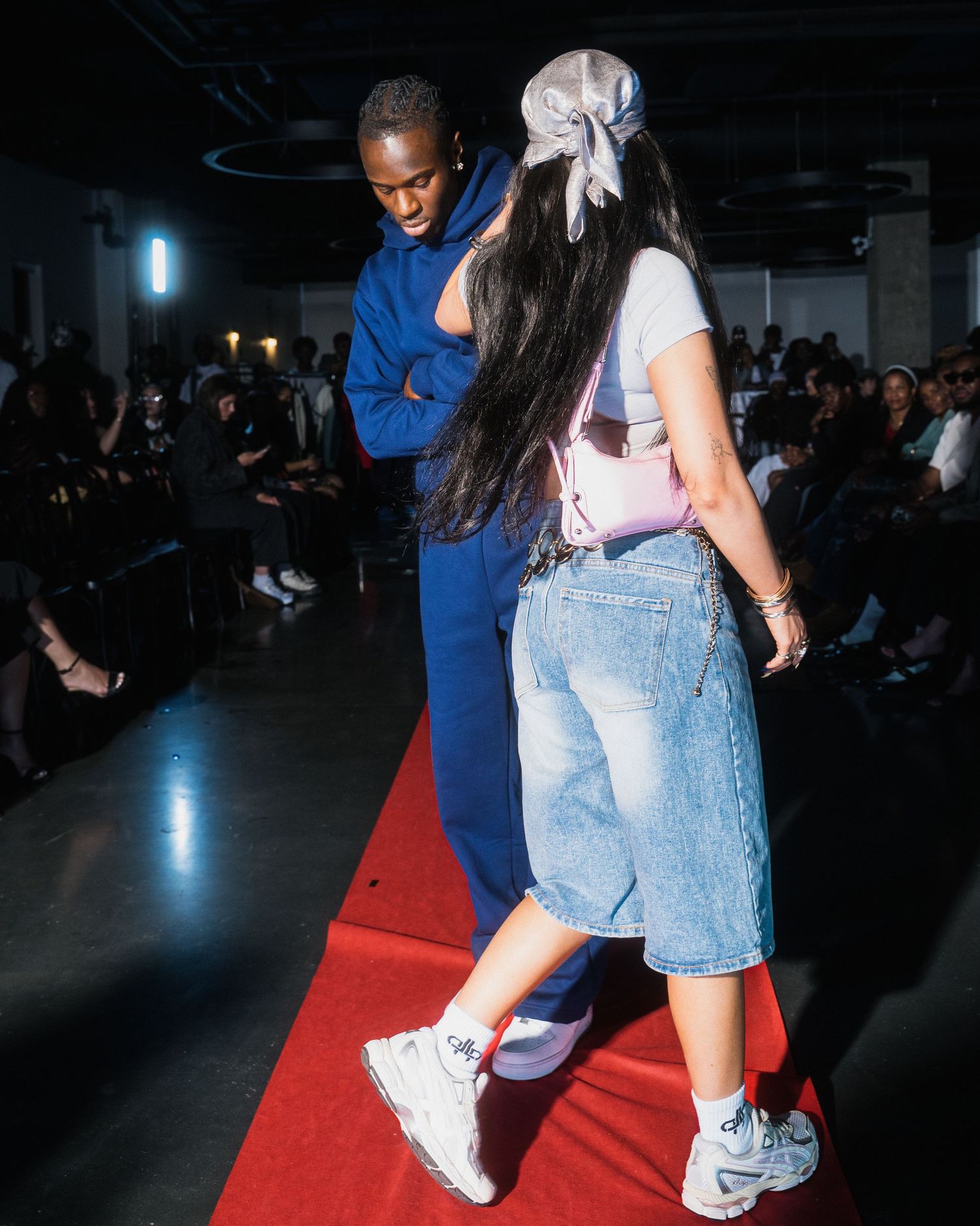 Two models walk a red carpet runway, one in blue jumpsuit and one in denim shorts and white sneakers.