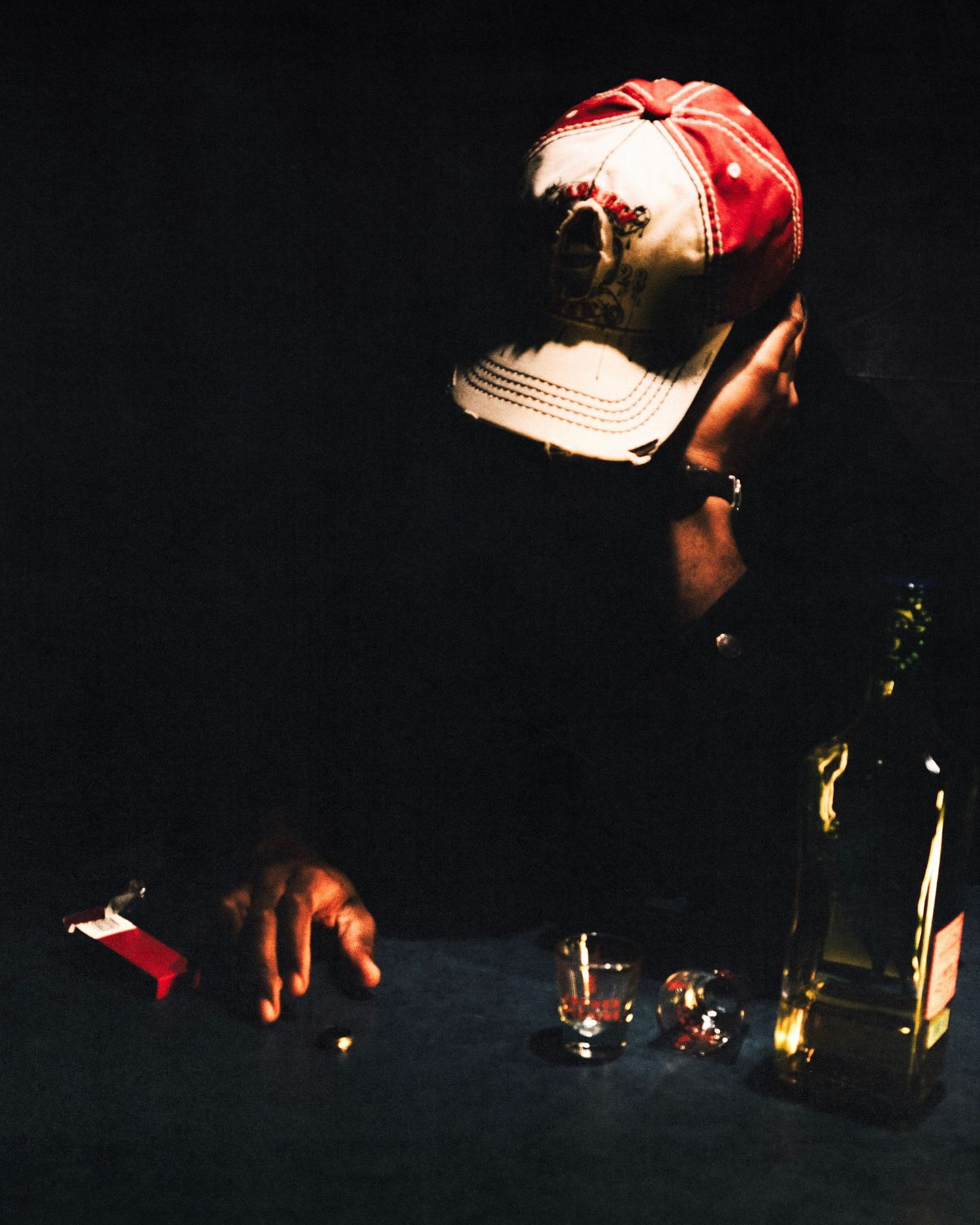 Person in red cap reaching toward bottles on dark table in dim lighting.