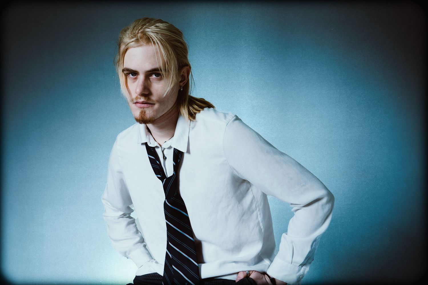 Young man with blonde hair in a white shirt and striped tie poses against a blue studio backdrop.