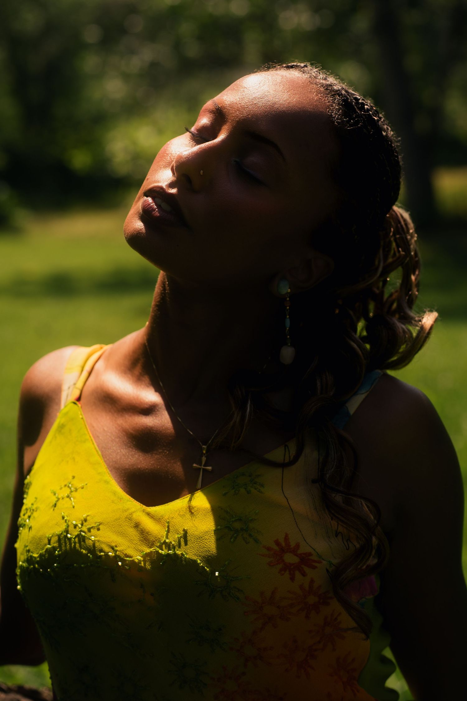 Woman in yellow floral dress with cross necklace tilting head back in sunlight outdoors.