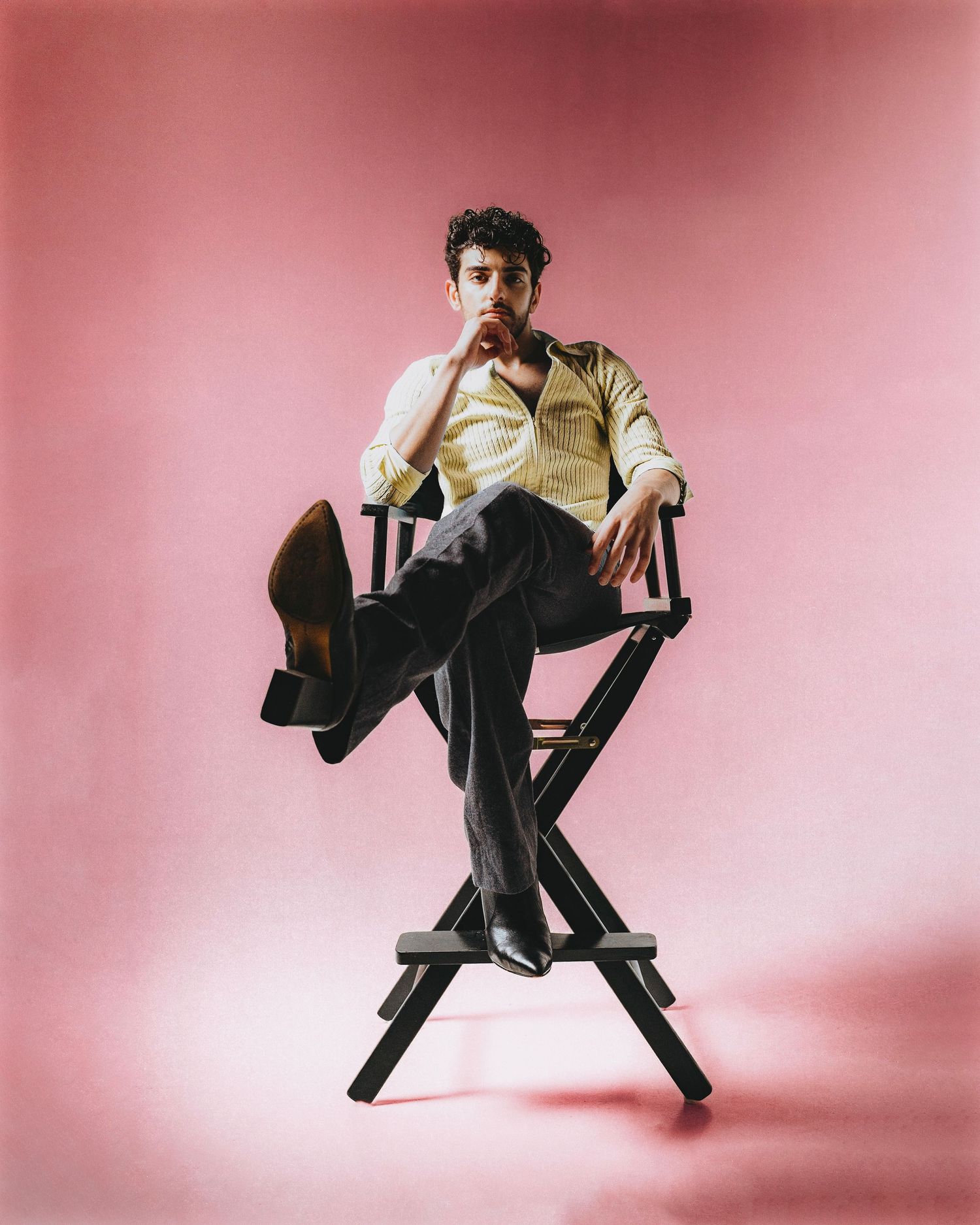 Man in yellow shirt and dark trousers poses on directors chair against pink background.