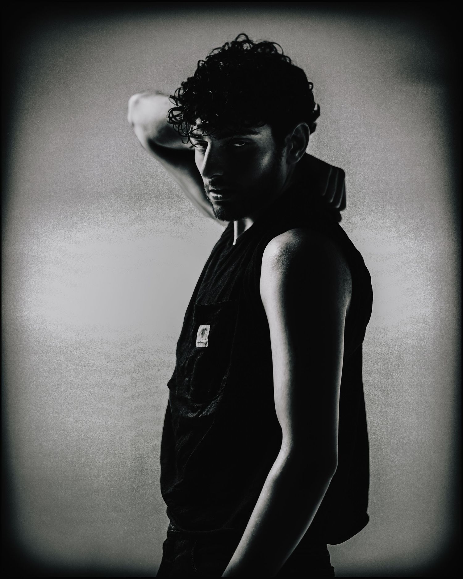 Black and white portrait of curly-haired man in sleeveless black hoodie looking over shoulder.