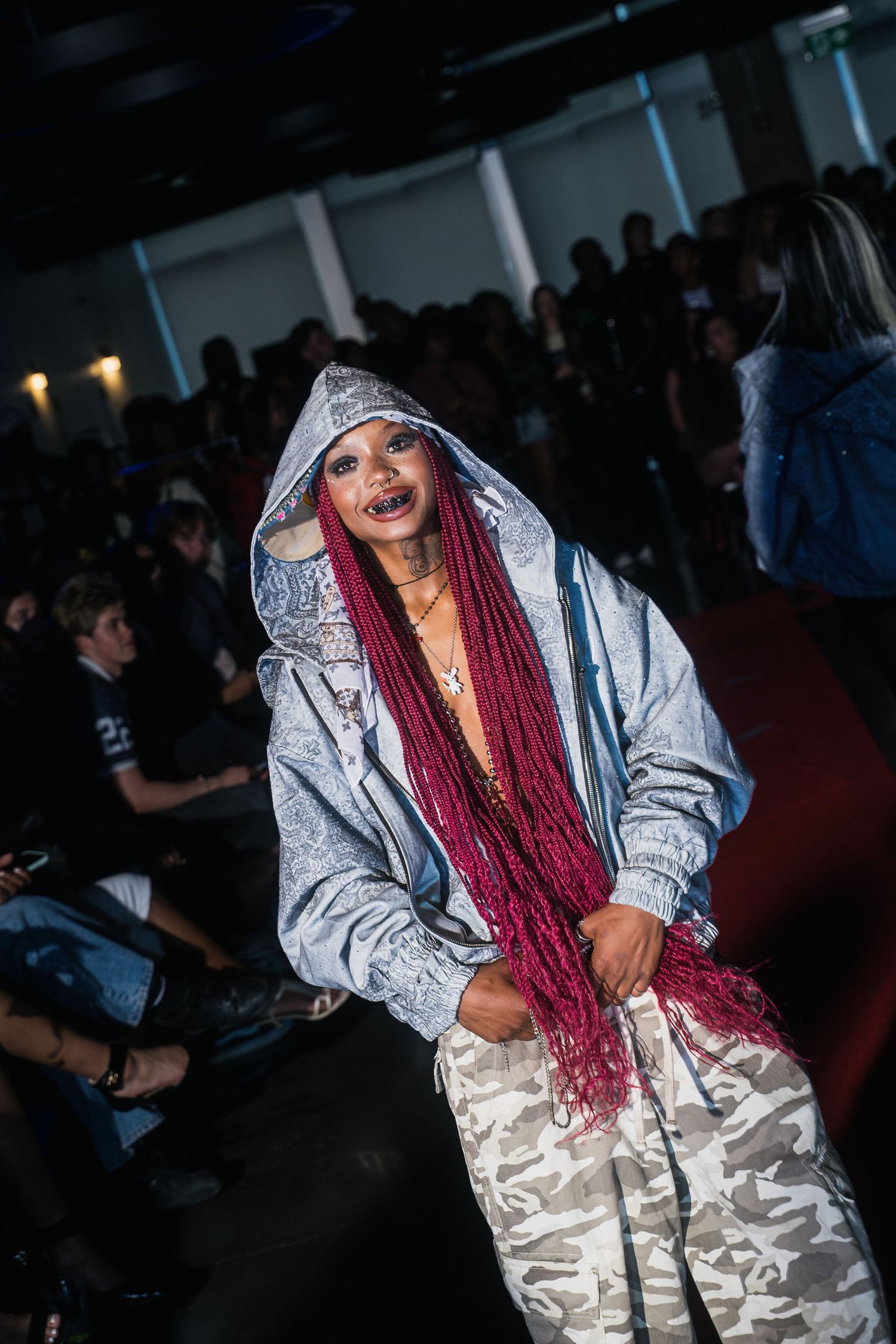 Stylish woman with burgundy box braids in a silver hoodie and camo pants posing on a red carpet runway.