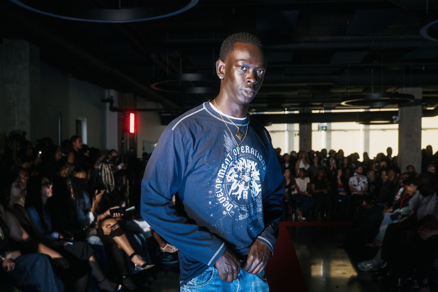 Male model poses on runway in navy graphic long sleeve shirt and light wash jeans.