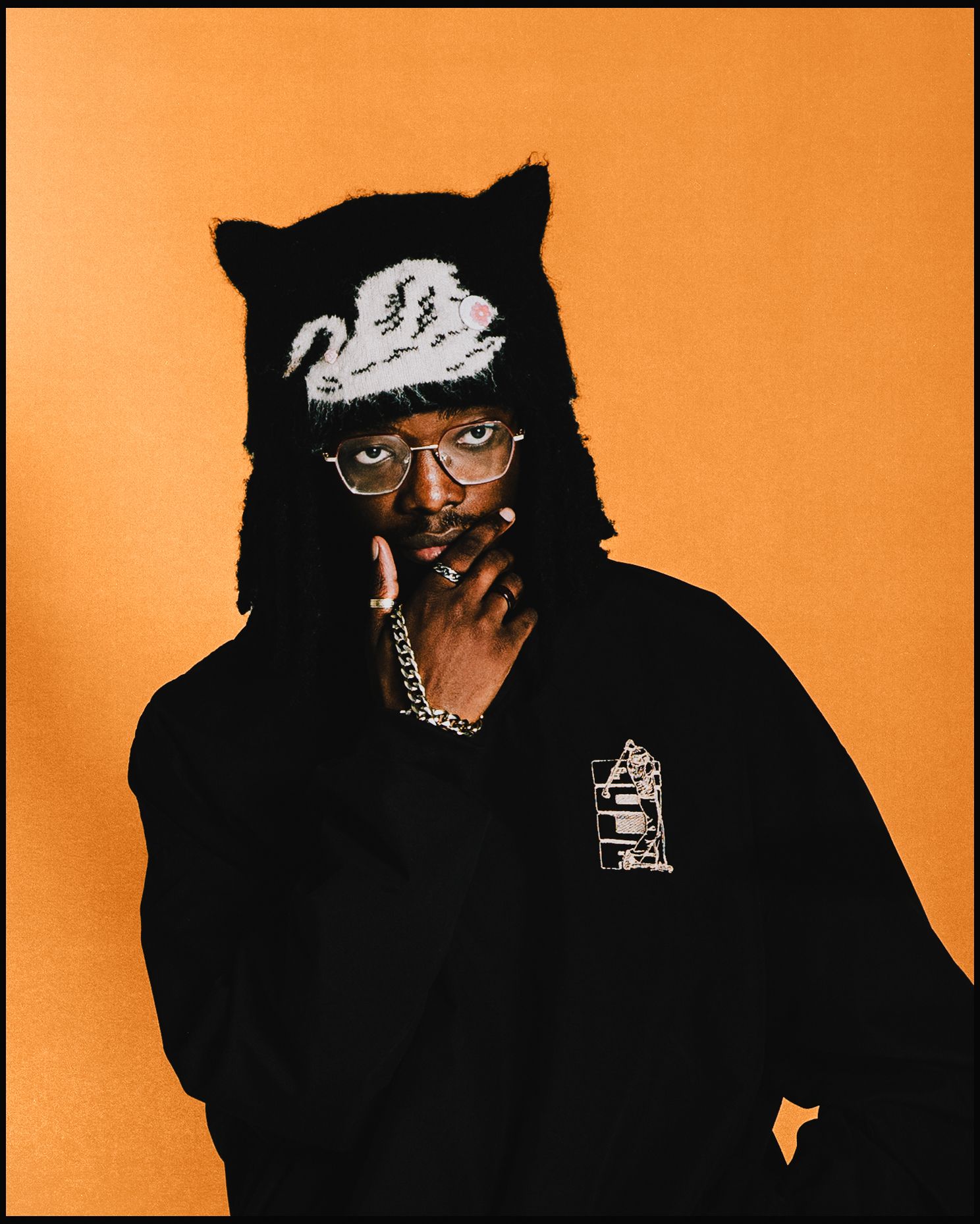 Artist poses in black outfit and cat-ear hat against orange background.