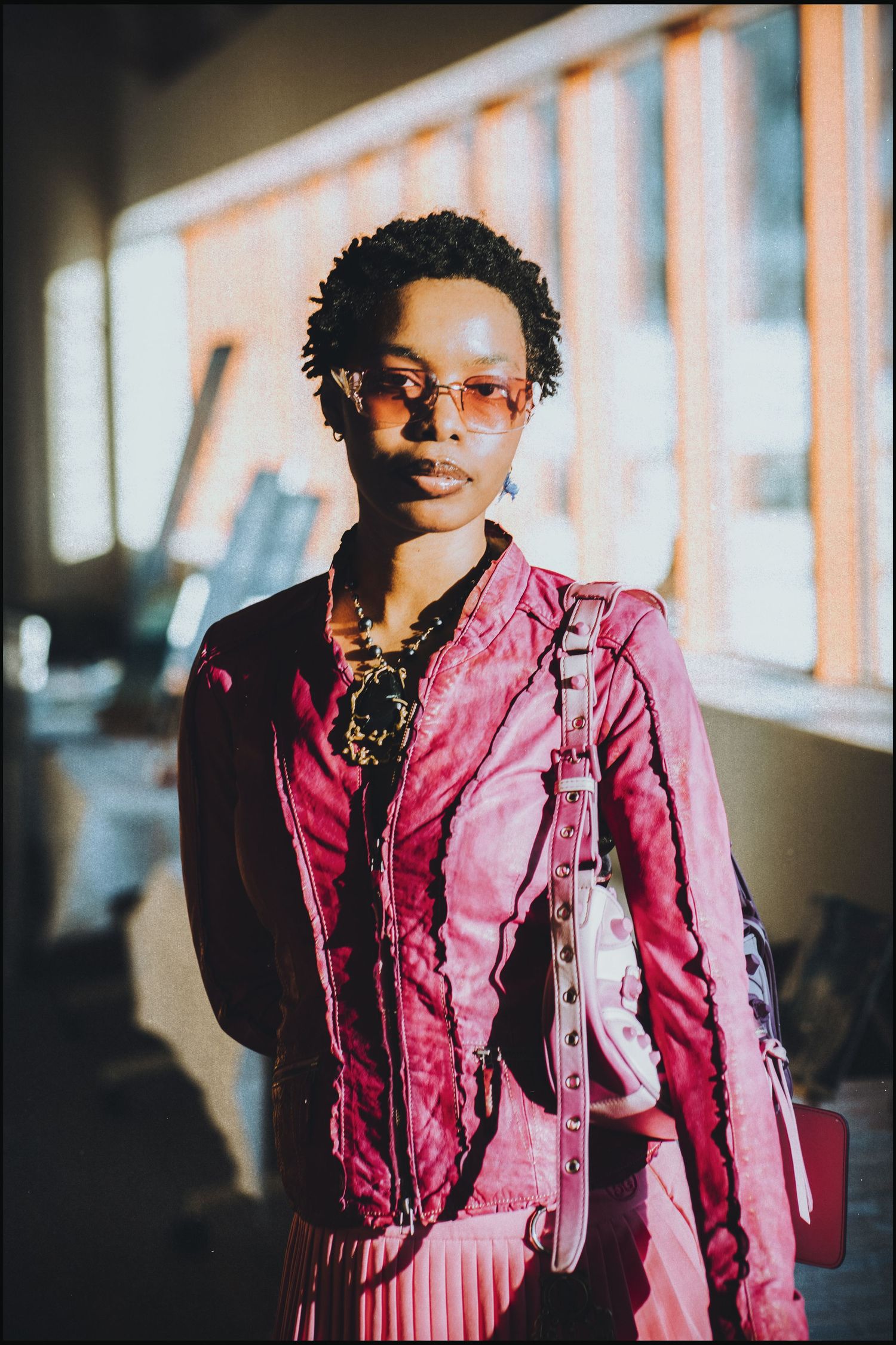 Stylish woman in pink jacket and tinted sunglasses poses indoors with pink bag.