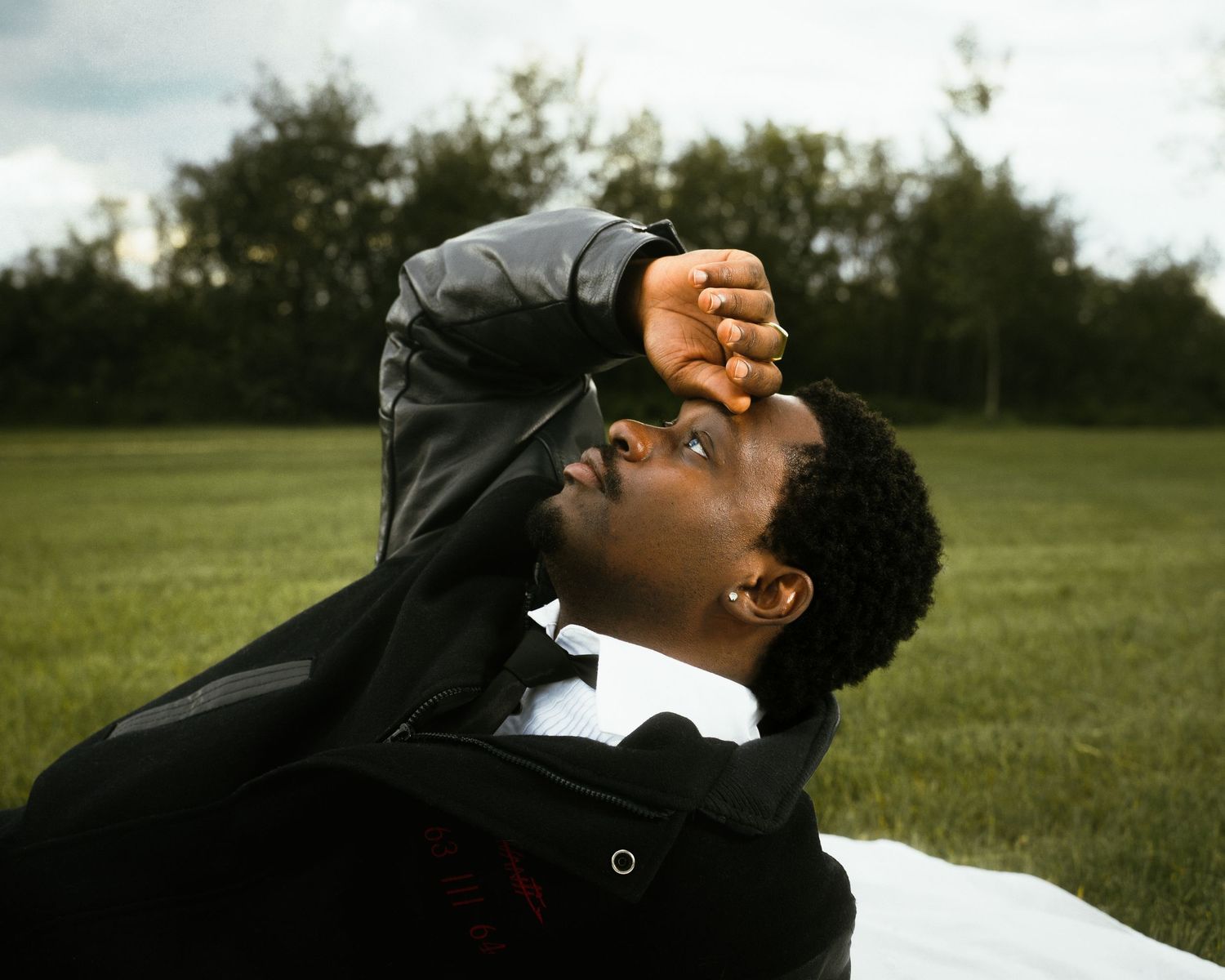 Man in black suit jacket looking up with fist raised, lying on green grass outdoors.