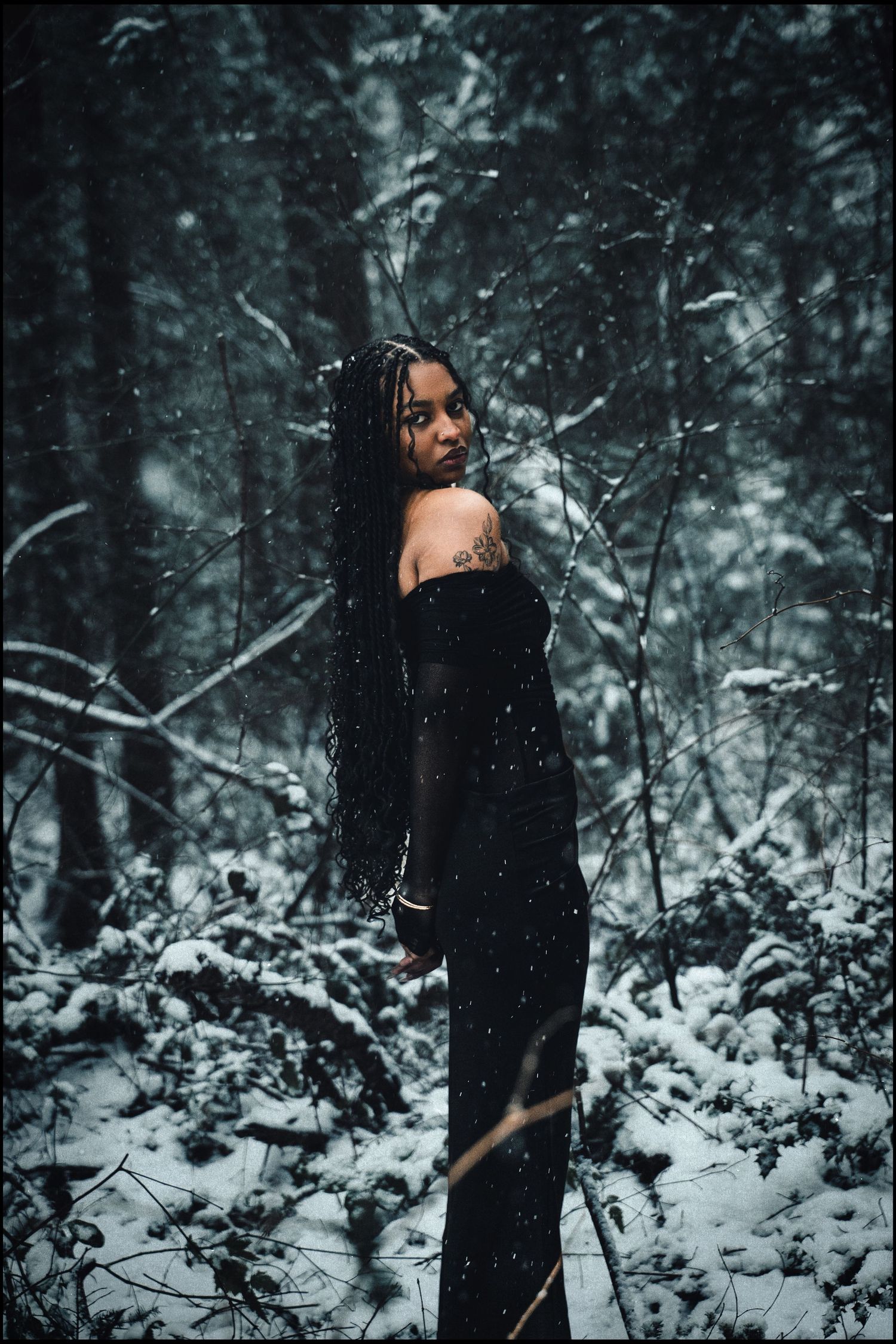 Woman in black dress with shoulder tattoo poses in snow-covered winter forest.