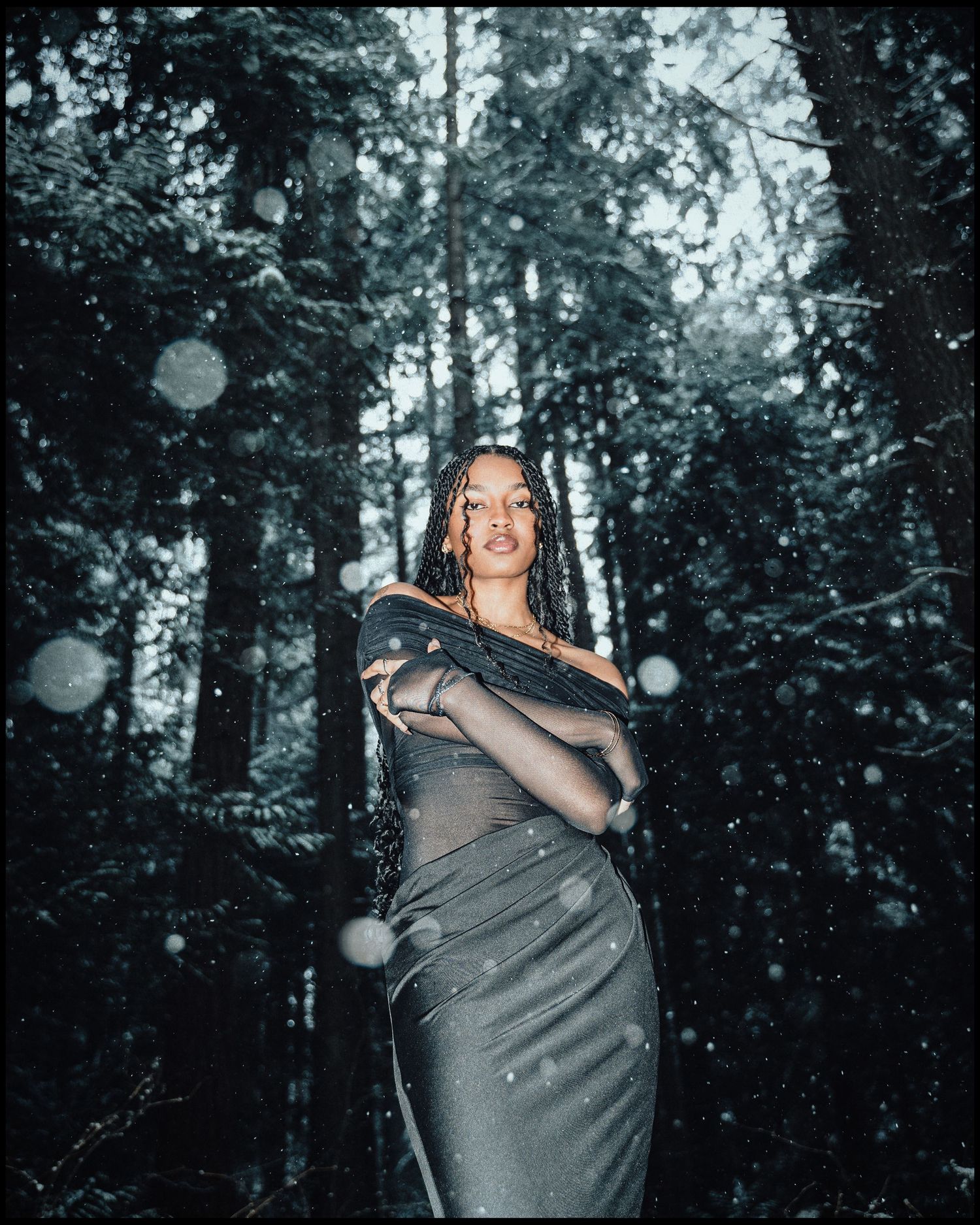 Woman in grey dress with arms crossed posing in snowy forest with bokeh snowflakes.