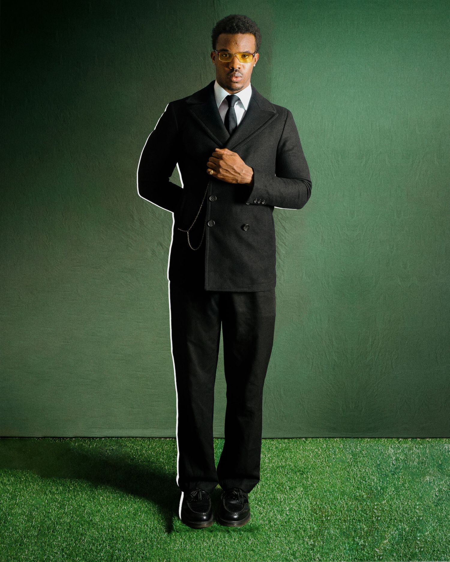 Man in black double-breasted suit and yellow sunglasses standing on green grass backdrop.
