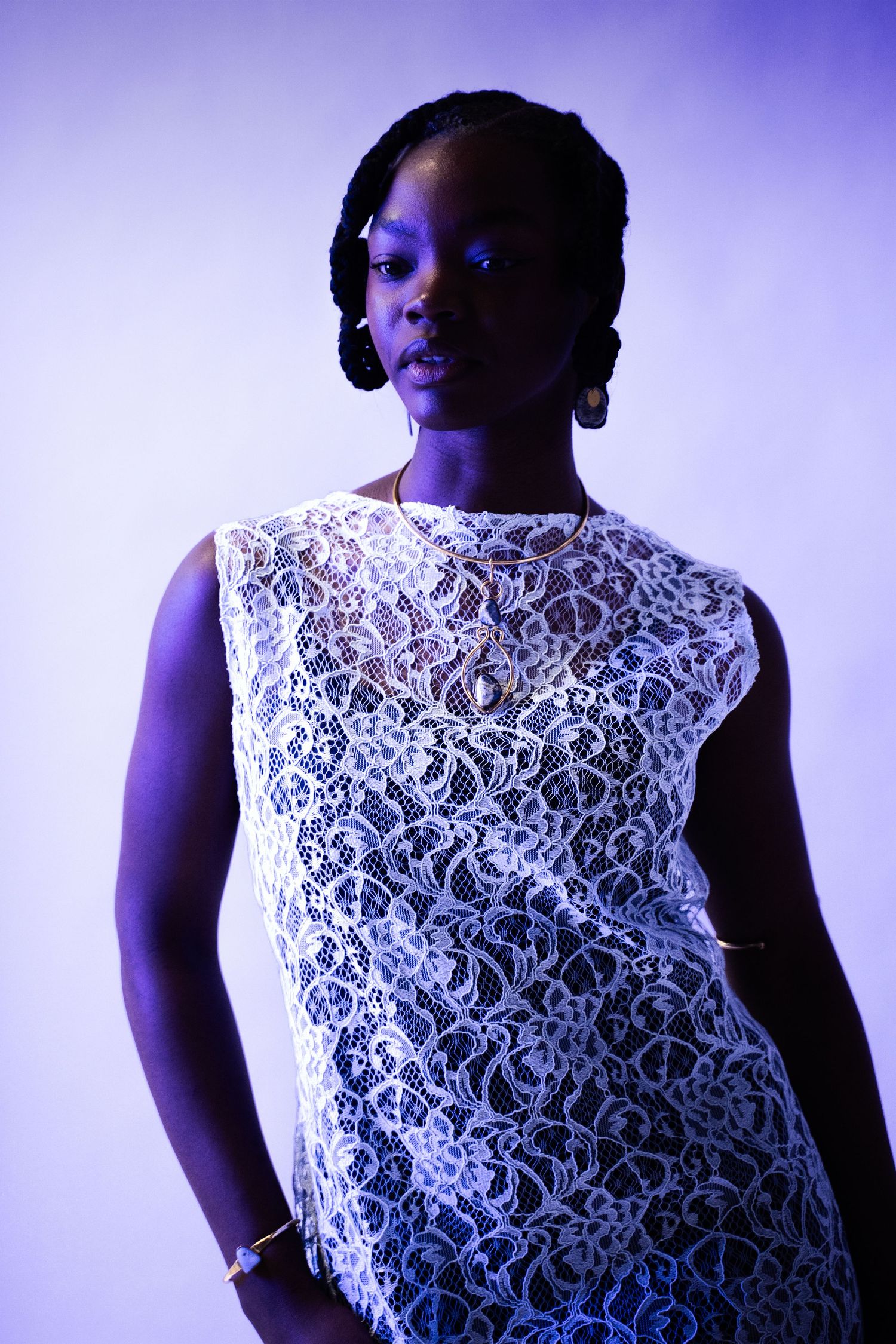 A young Black woman poses in a white lace sleeveless dress under purple studio lighting.