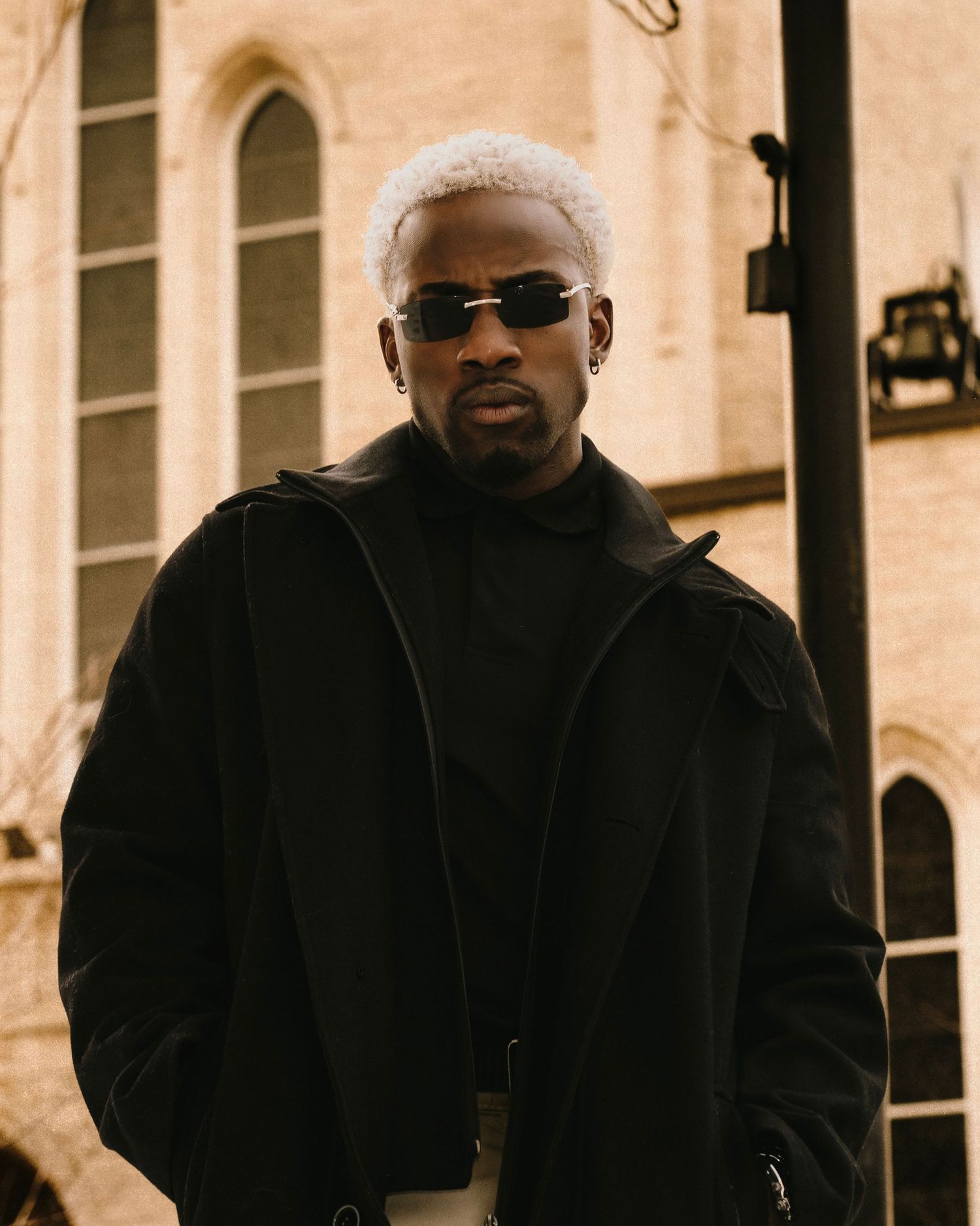 Fashionable man with platinum hair wearing all-black outfit and sunglasses poses before historic church.