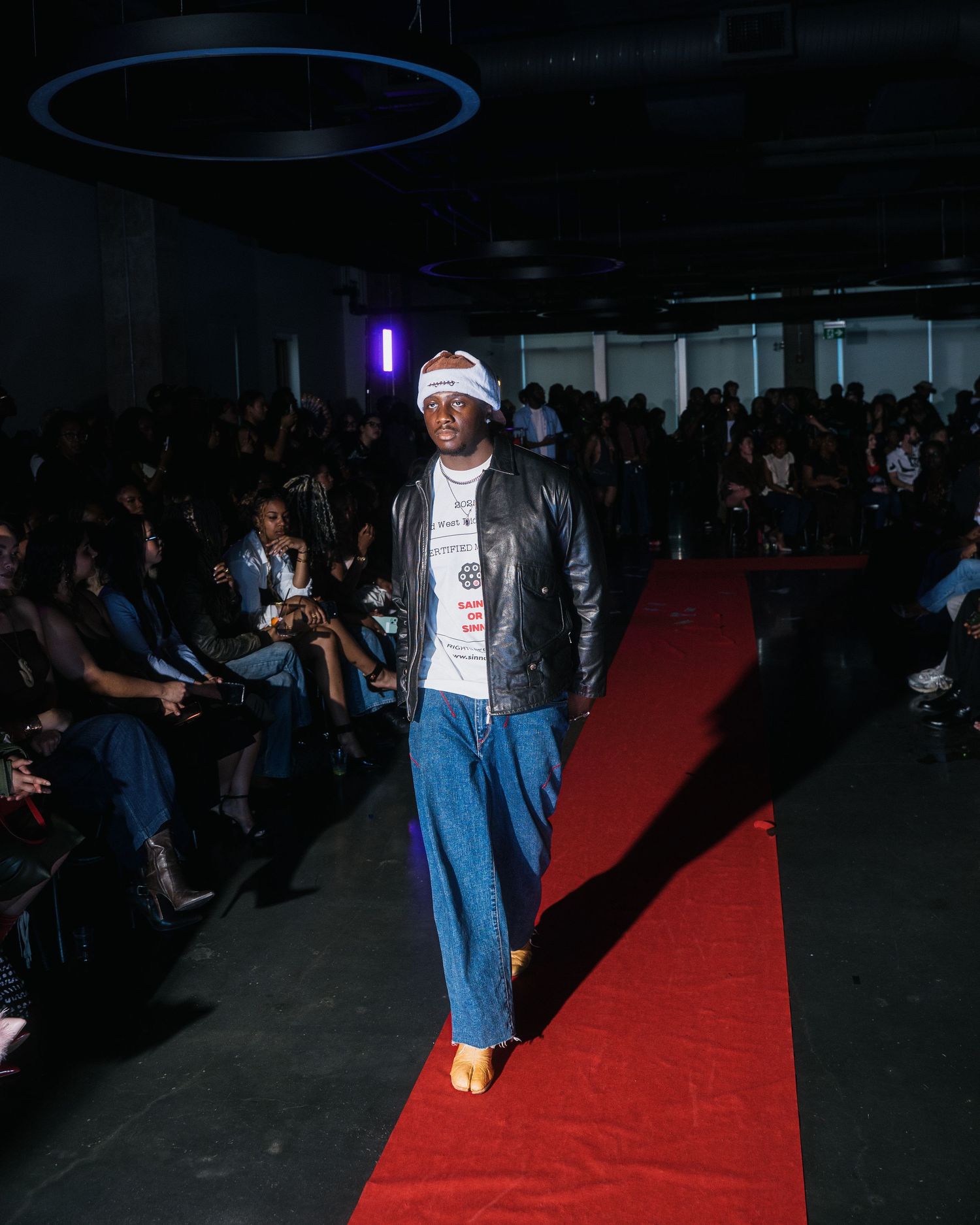 Male model walks red carpet runway wearing leather jacket, white graphic tee, wide-leg jeans, and white beanie.