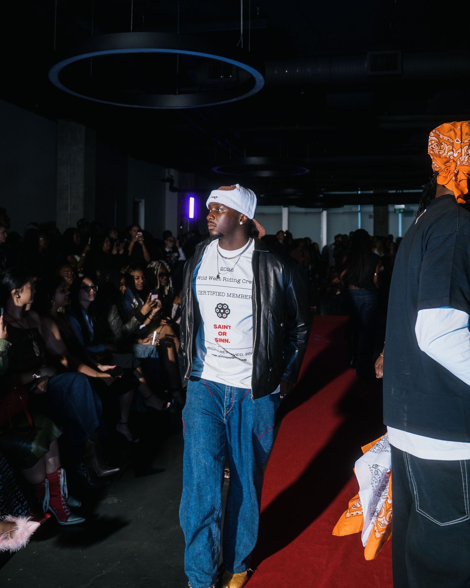 Male model stands on red carpet runway in leather jacket, white graphic tee, and wide-leg denim jeans.