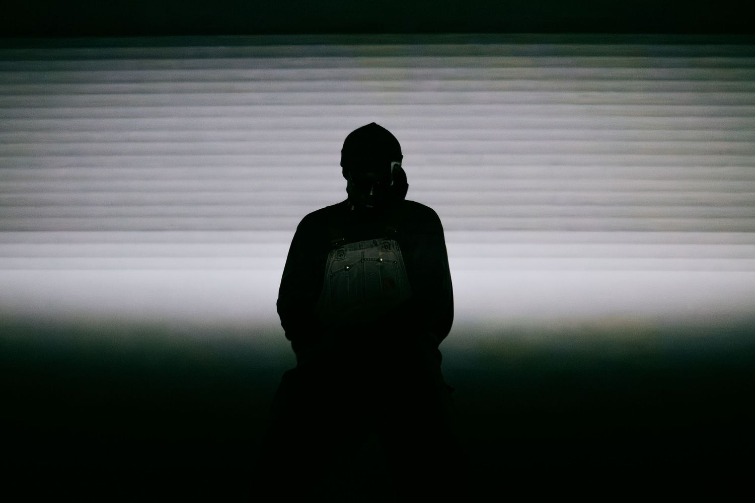 Silhouette of a person standing alone in front of illuminated horizontal blinds in a dark room.