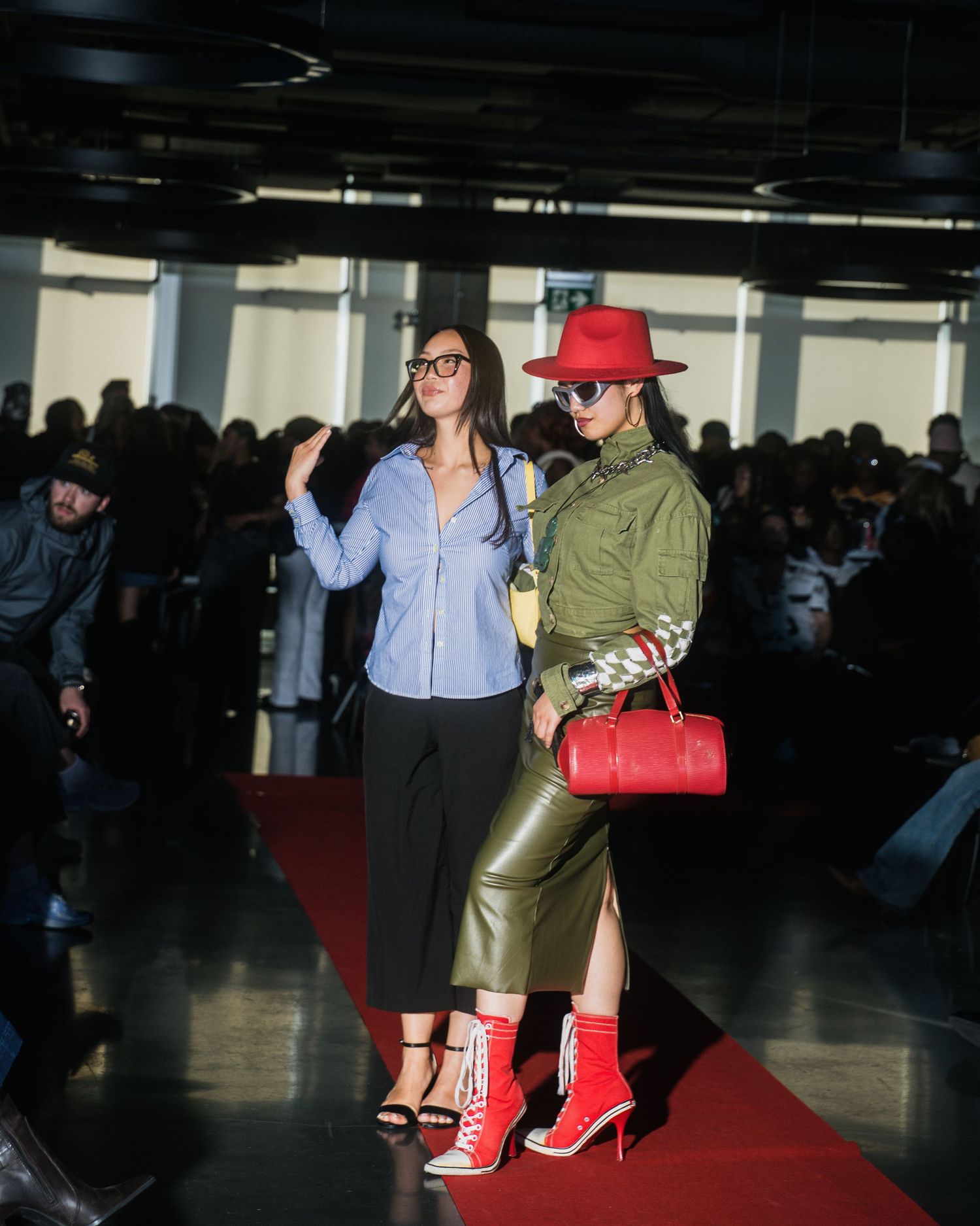 Two models on red carpet runway, one wearing red hat, olive jacket, leather skirt and red boots.