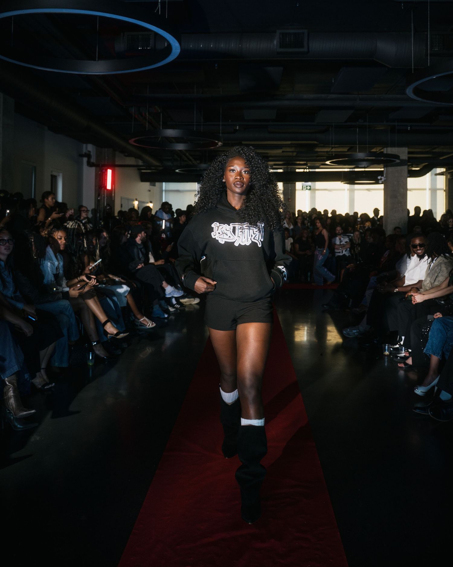 Female model walks runway in black graphic hoodie and shorts with knee-high boots.