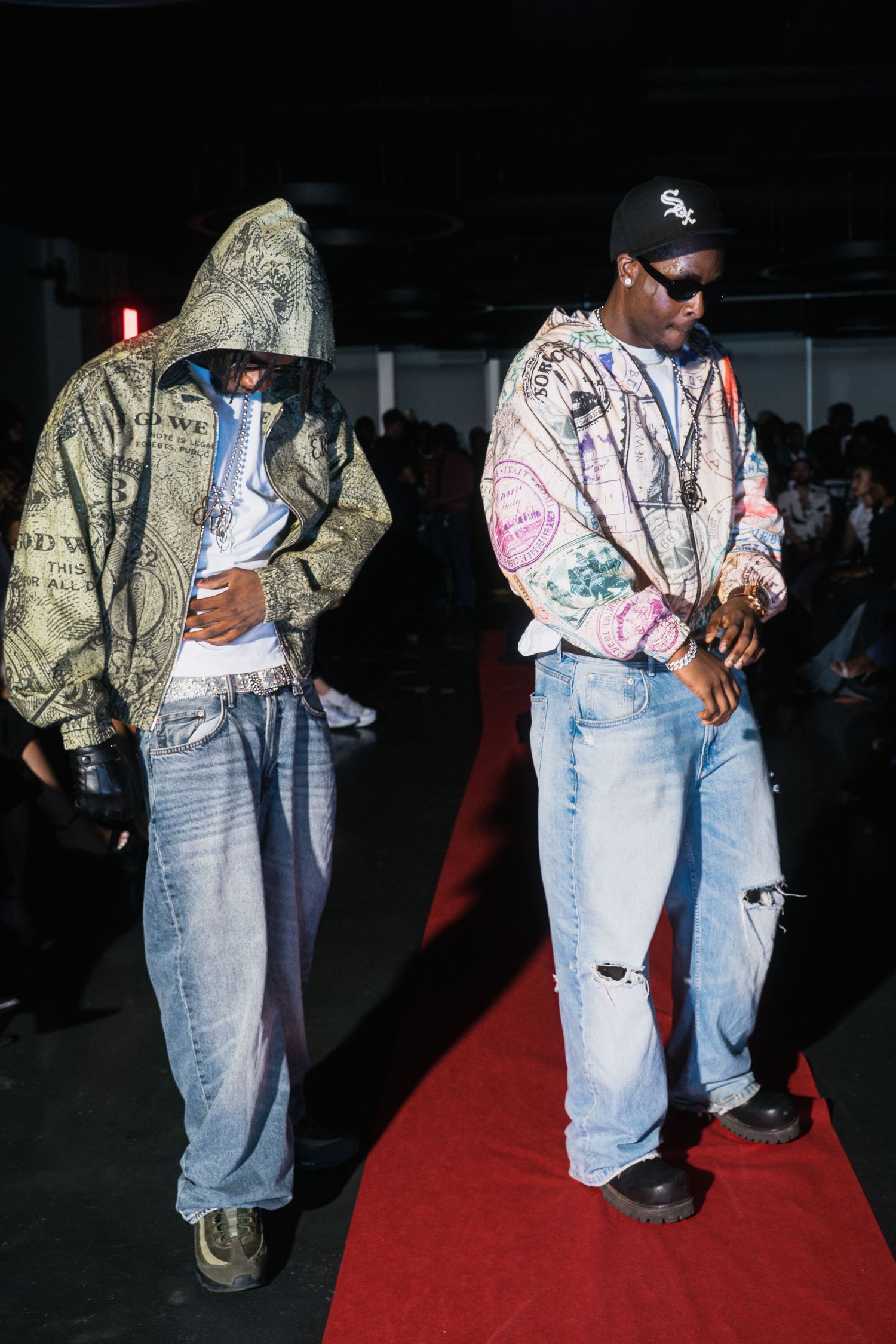 Two men in streetwear hoodies and baggy jeans walking a red carpet at an urban fashion show.