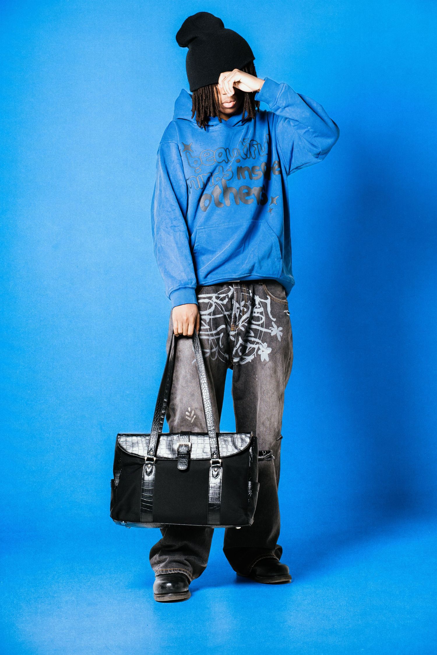Person in blue graffiti hoodie, black beanie, and distressed jeans holding a black leather bag against blue background.
