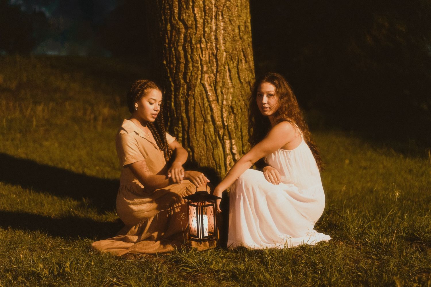 Two women in elegant dresses sit beside a glowing lantern under a large tree in golden evening light.