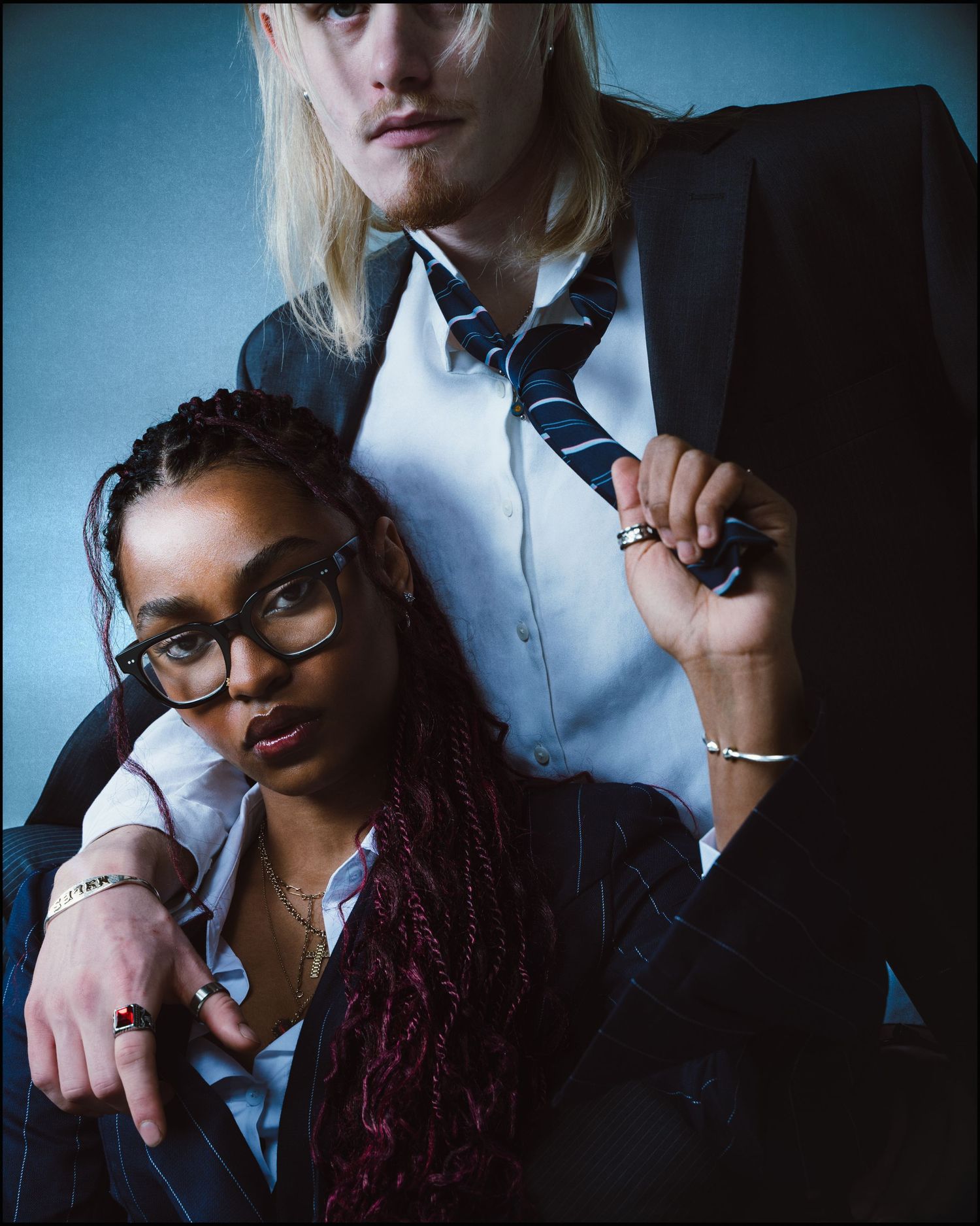 Close-up of a Black woman with braids and glasses leaning against a tall blonde man in a blazer.