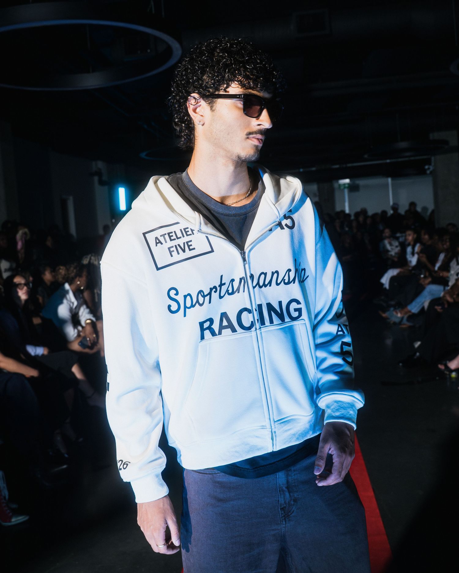 Male model in Atelier Five Sportsmanship Racing white zip hoodie walking a fashion show runway.