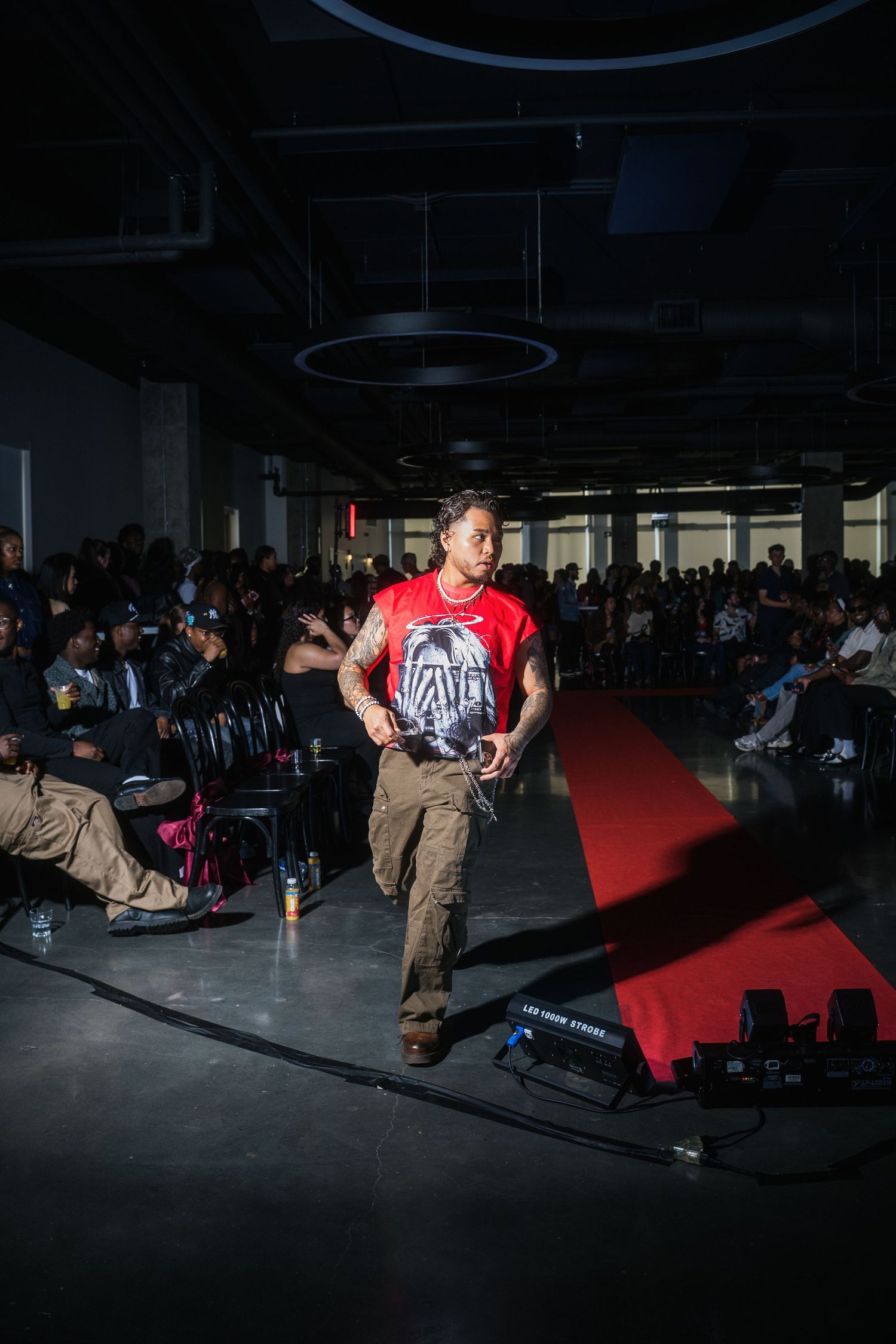 Tattooed model in red graphic tee and cargo pants walks red carpet runway at fashion show.